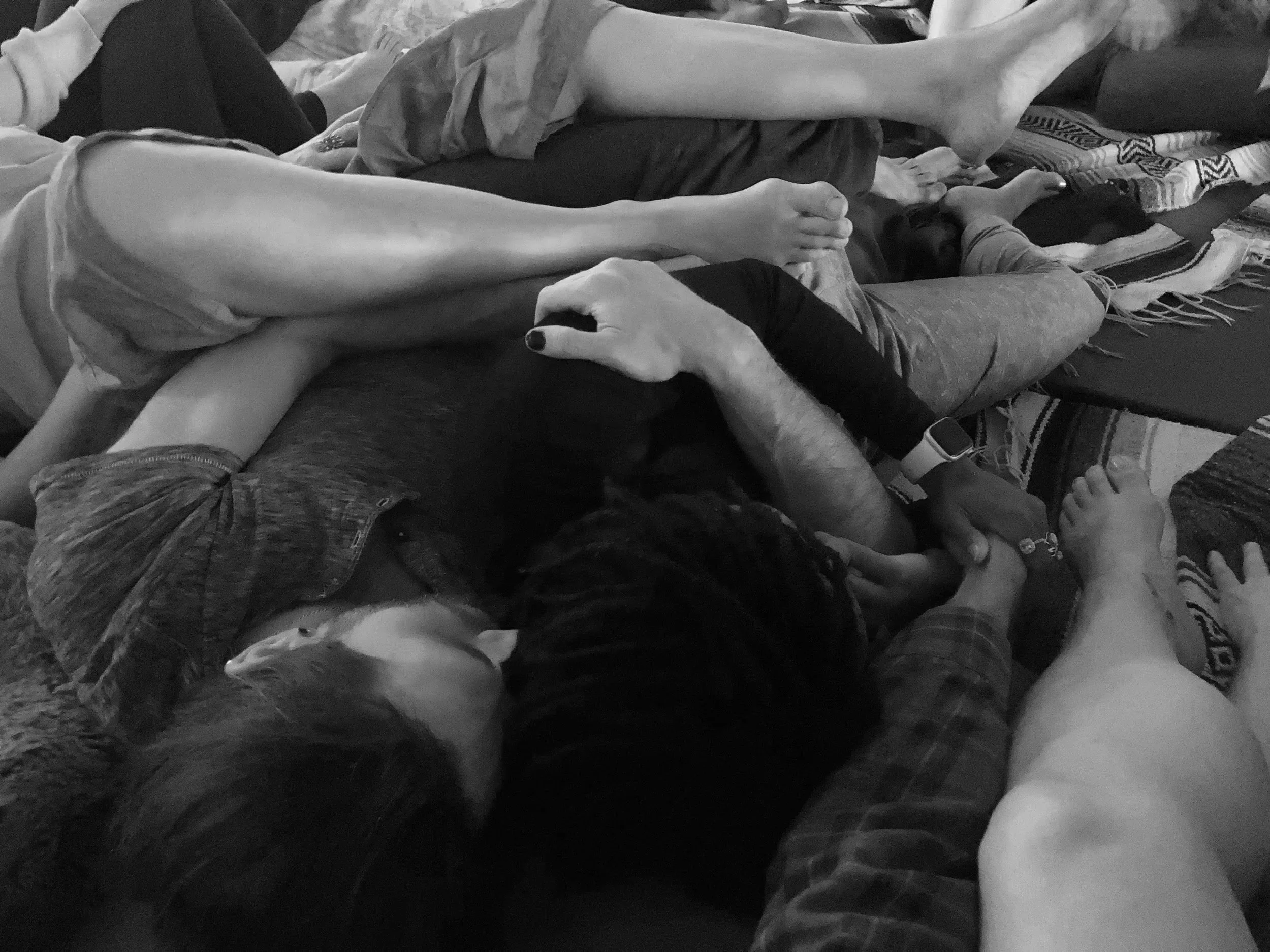 People lying down closely together, some with their arms intertwined, on a variety of blankets and mats, possibly relaxing or resting.
