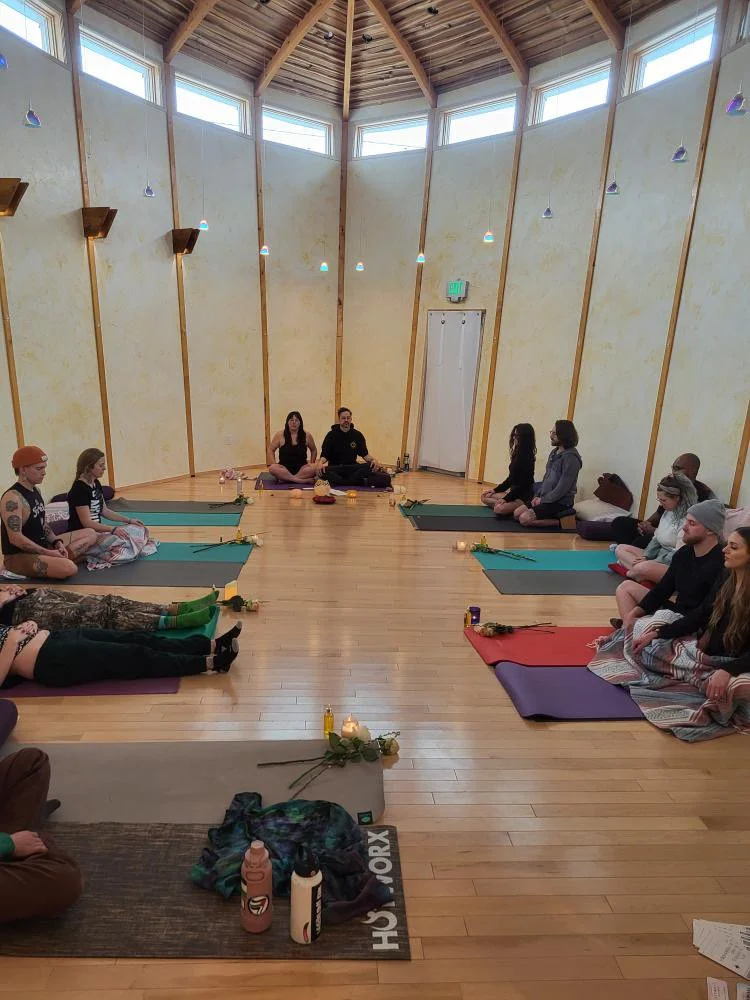 A group of people seated cross-legged on yoga mats in a circle inside a wooden-floored, high-ceiling room with large windows and soft lighting, participating in a meditation or yoga class.