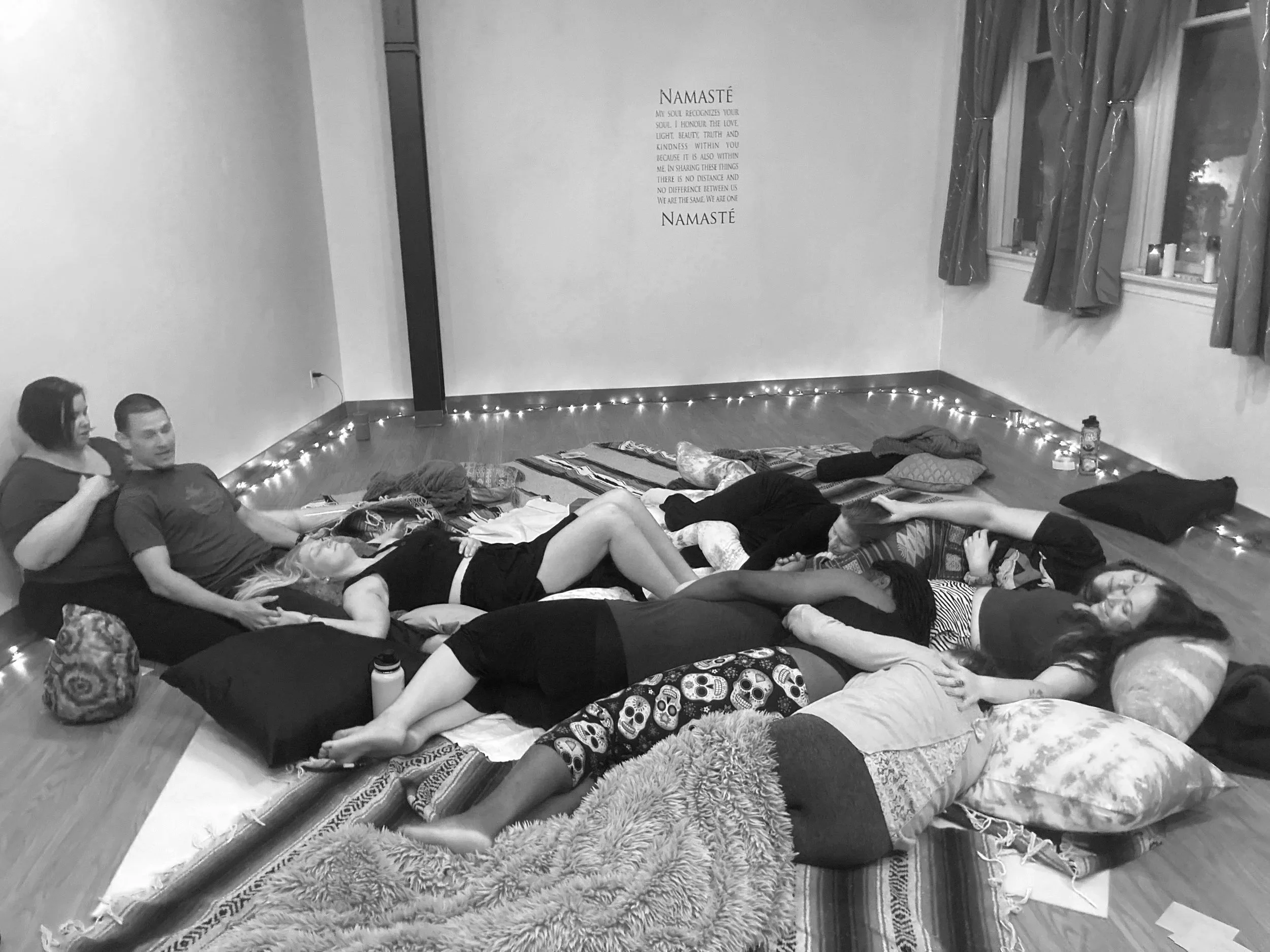 People lying on the floor with mats, blankets, and pillows in a circle, likely at a yoga or meditation session, with string lights along the wall and a namaste quote on the wall.