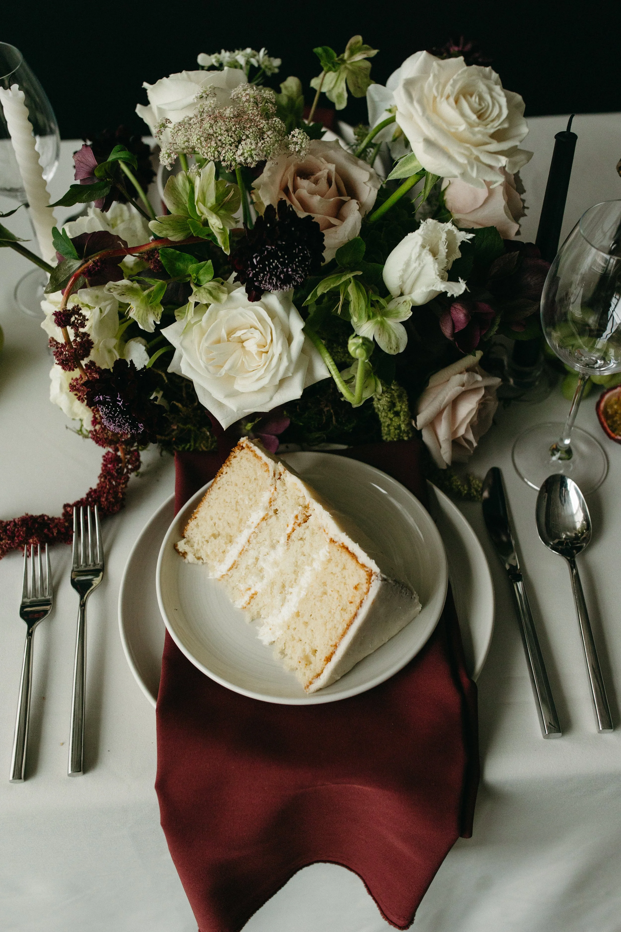 A table setting featuring a small white wedding cake slice on a reception table with burgundy and green flowers by Petals & Loam Calgary wedding florist.