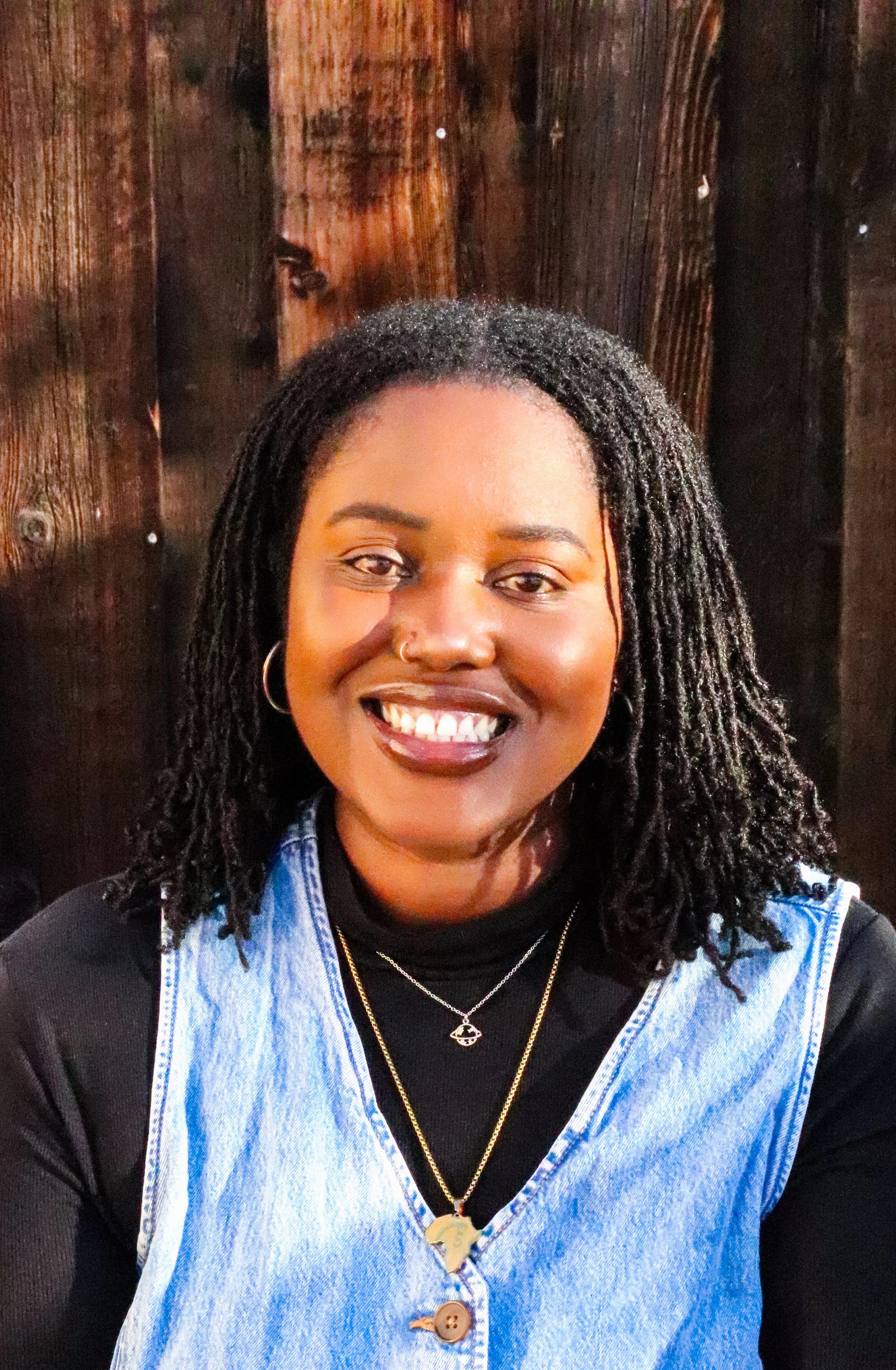 A picture of Bayana A. Davis, a Black woman with dark brown skin and shoulder-length microlocs. She's wearing a black turtleneck, denim vest, and gold jewelry. She's sitting in front of a wooden background.