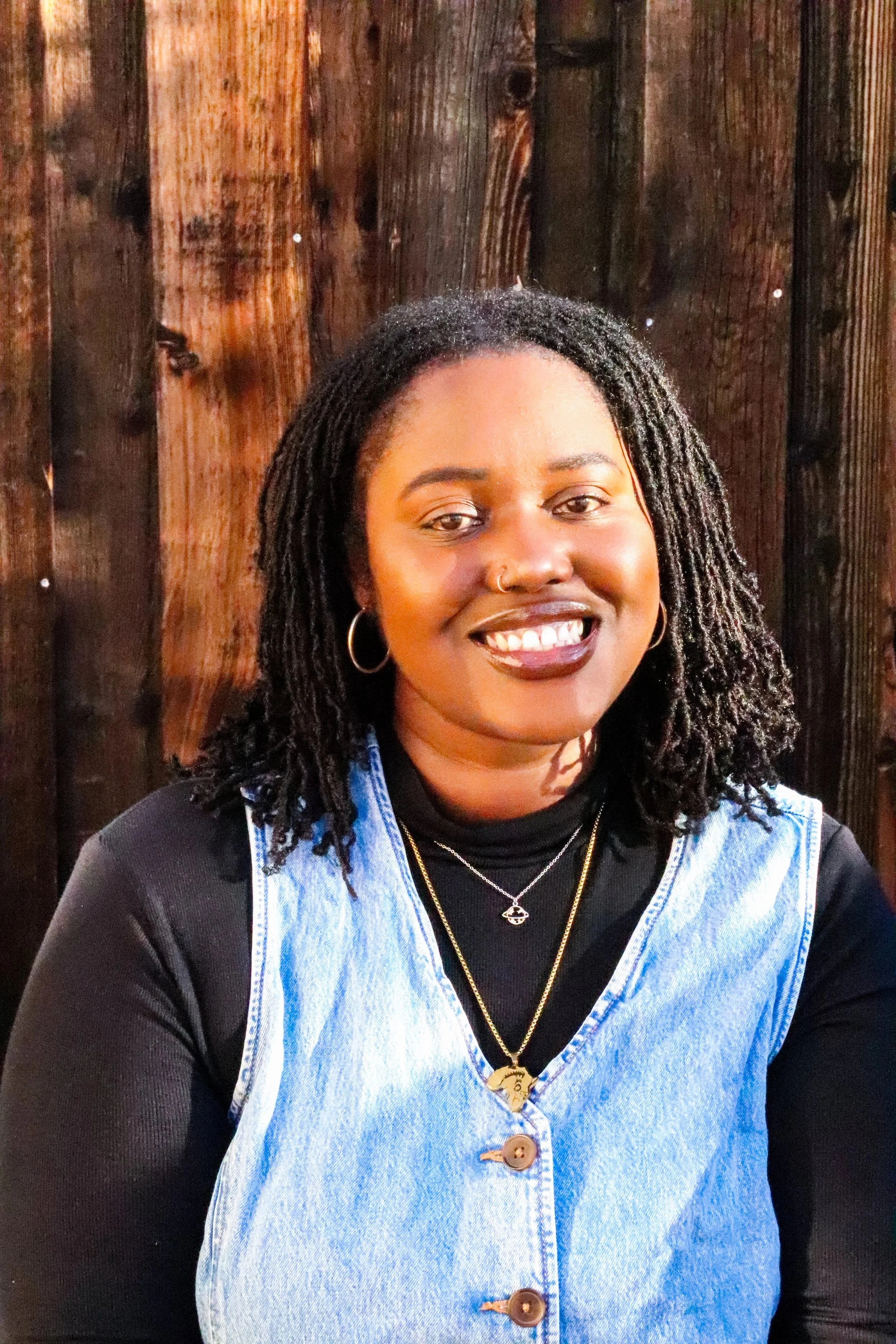 A picture of Bayana A. Davis, a Black woman with dark brown skin and shoulder-length microlocs. She's wearing a black turtleneck, denim vest, and gold jewelry. She's sitting in front of a wooden background.