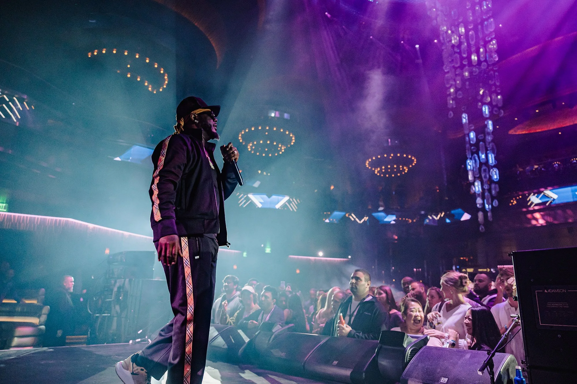 A male performer in black clothing, sunglasses, and a cap holds a microphone on stage during a concert with colorful lights and a lively audience in a large indoor venue.