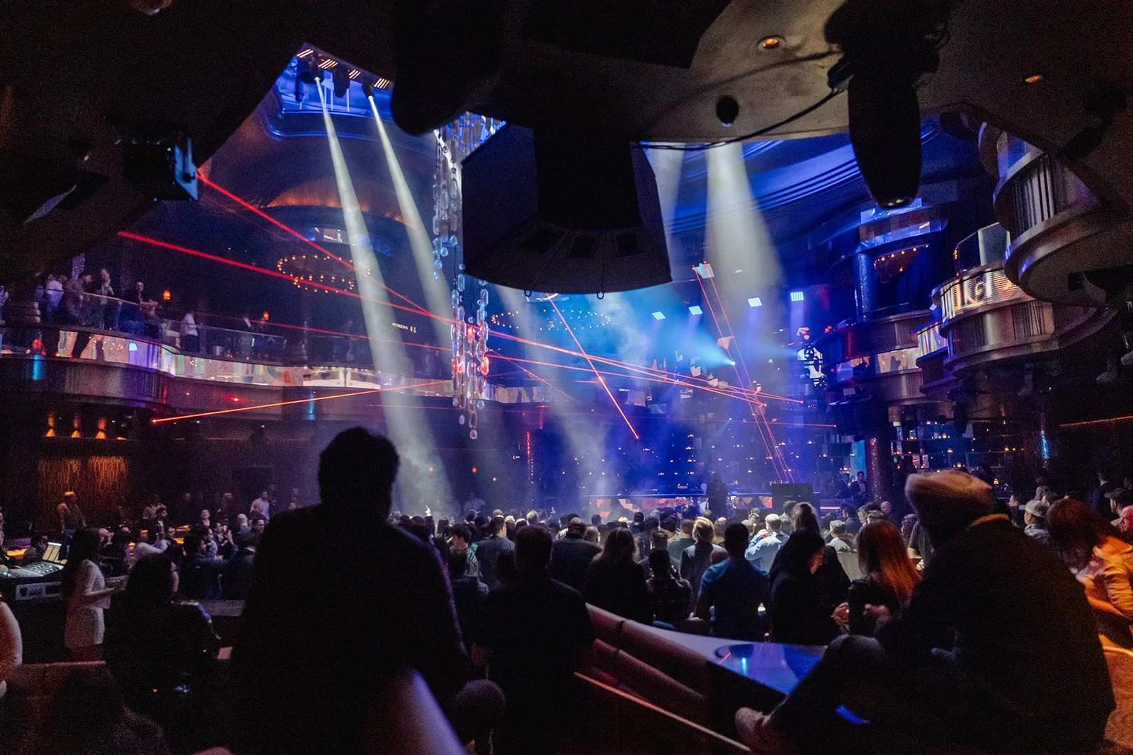 An indoor concert venue with a stage, colorful lighting, and laser effects. Audience members are seated and standing, watching a performance, with balconies overlooking the main floor.