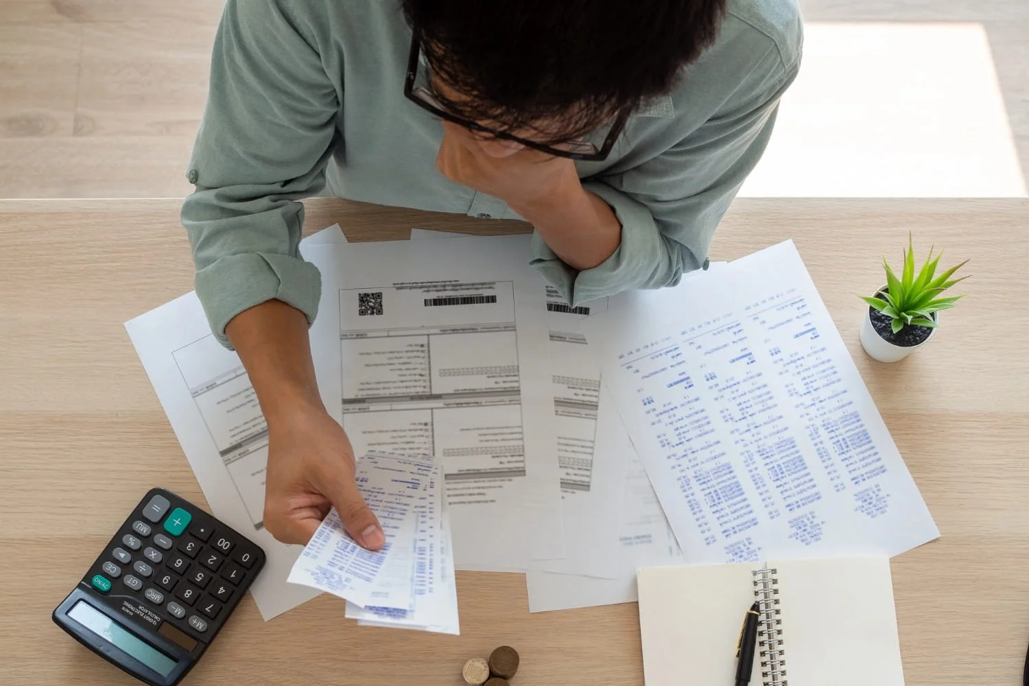 A person wearing glasses seated at a desk with a calculator, a notebook, and a pen, reviewing receipts and bills.