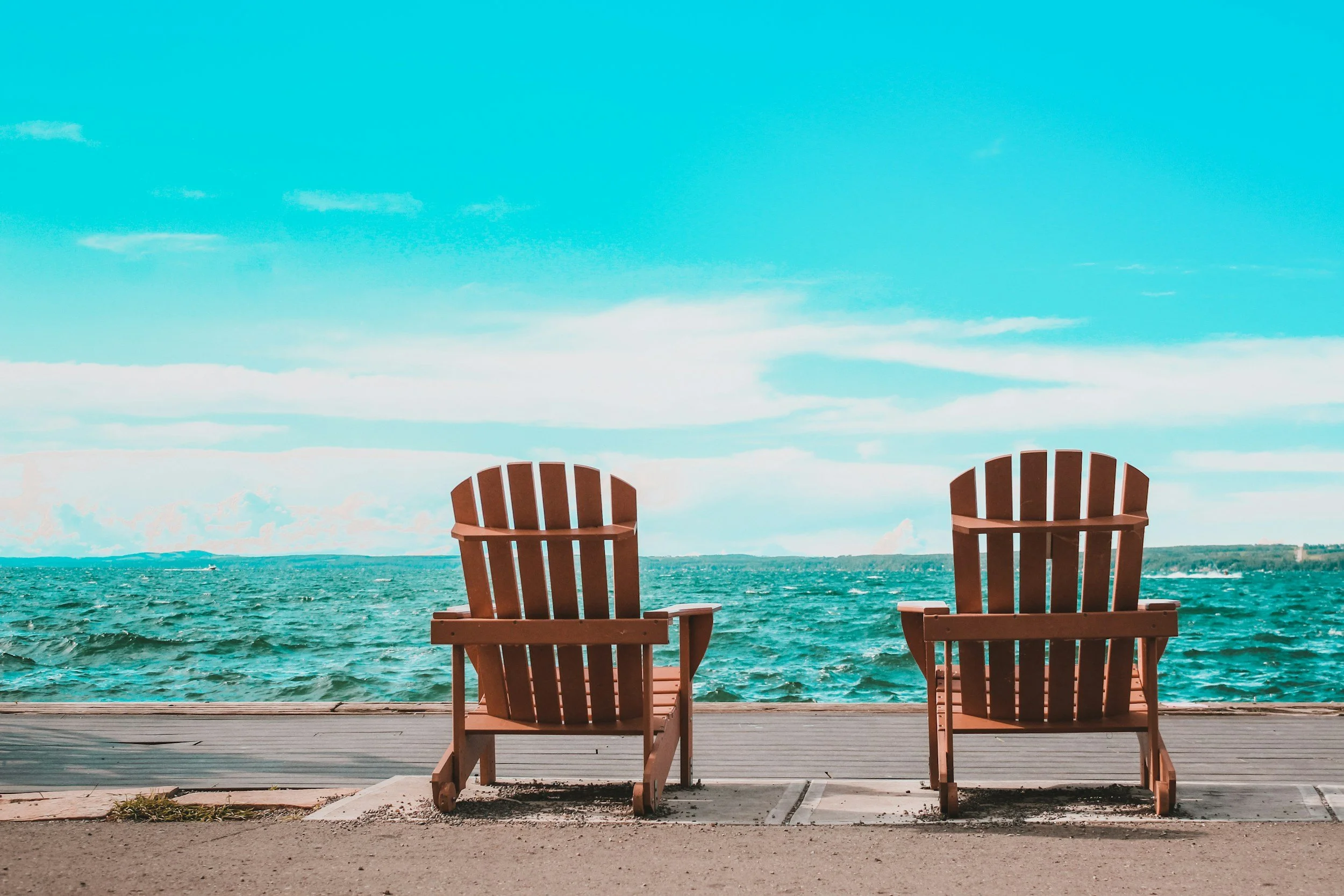 Two wooden Adirondack chairs on a beach boardwalk facing the ocean, under a partly cloudy sky.