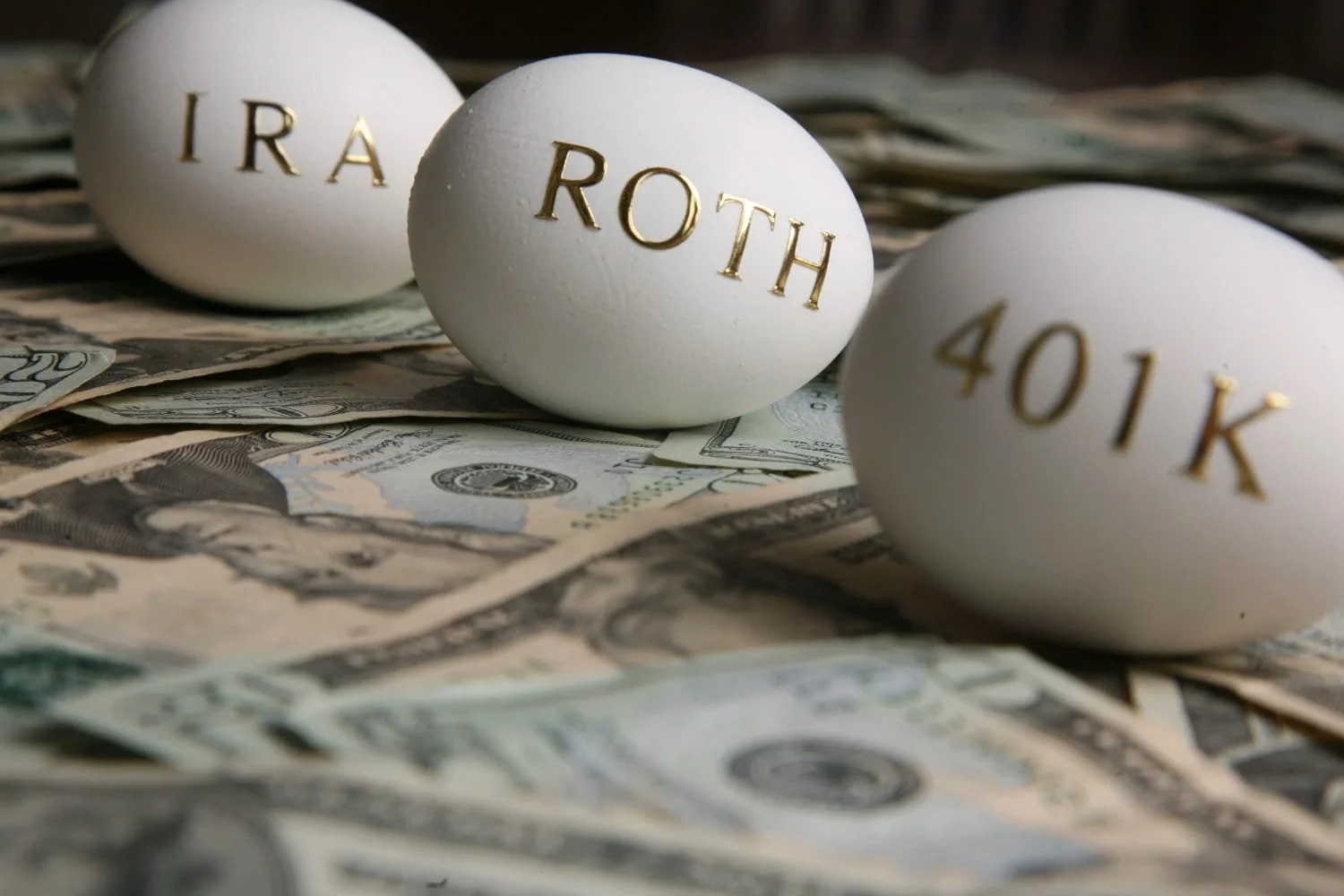 Three white eggs with IRA, ROTH, and 401K written on them as they lay on a pile of money.