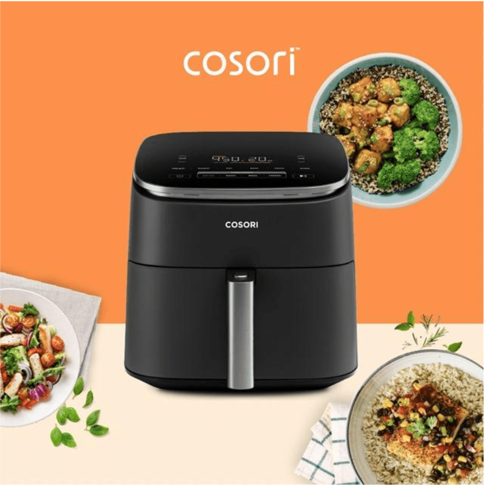 Black Cosori air fryer on a table with bowls of various cooked dishes surrounding it, set against a two-tone orange and beige background.