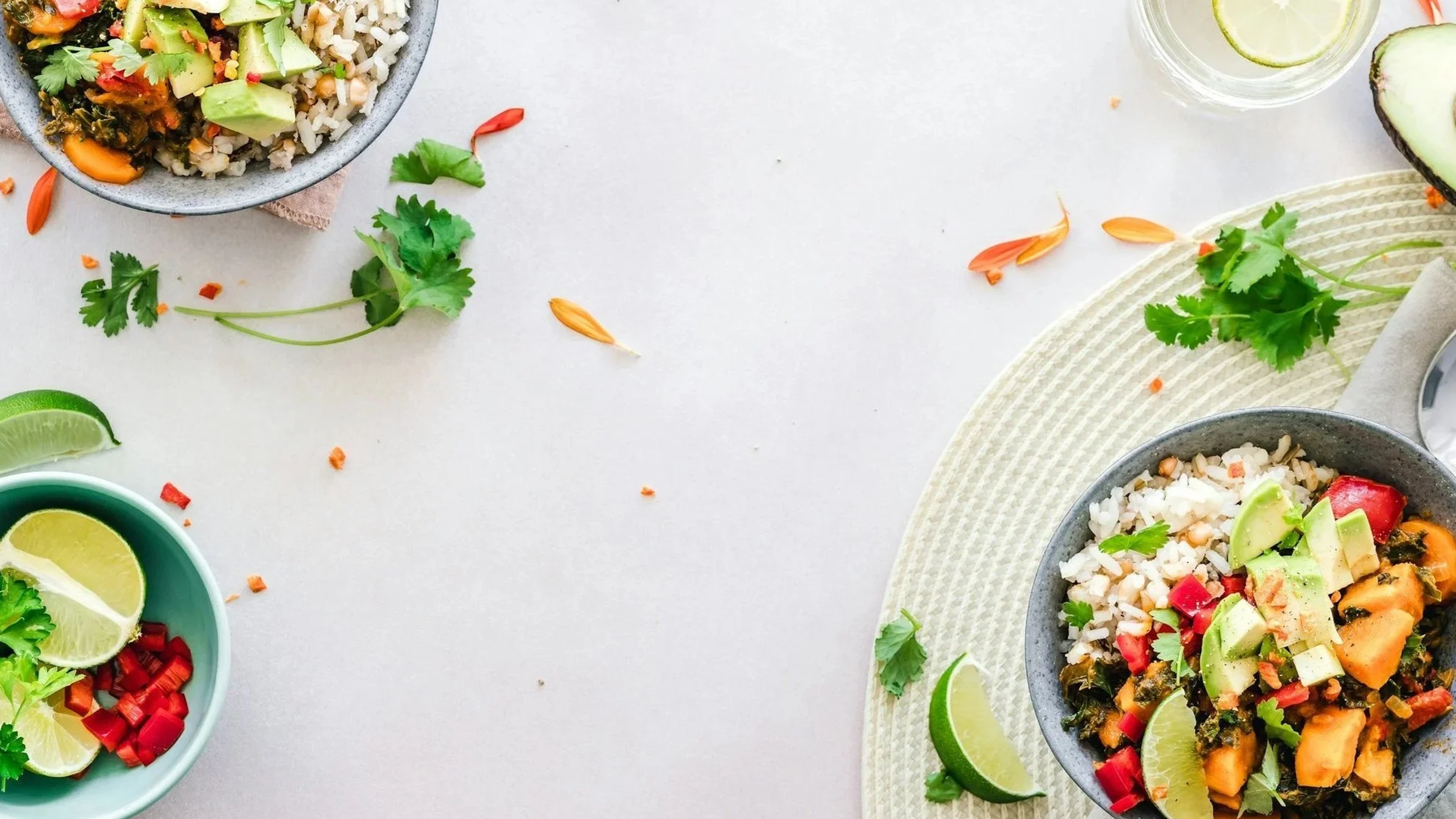 Bowls of rice and vegetables, lime wedges, cilantro, and sliced red peppers on a white surface with scattered cilantro leaves and flower petals