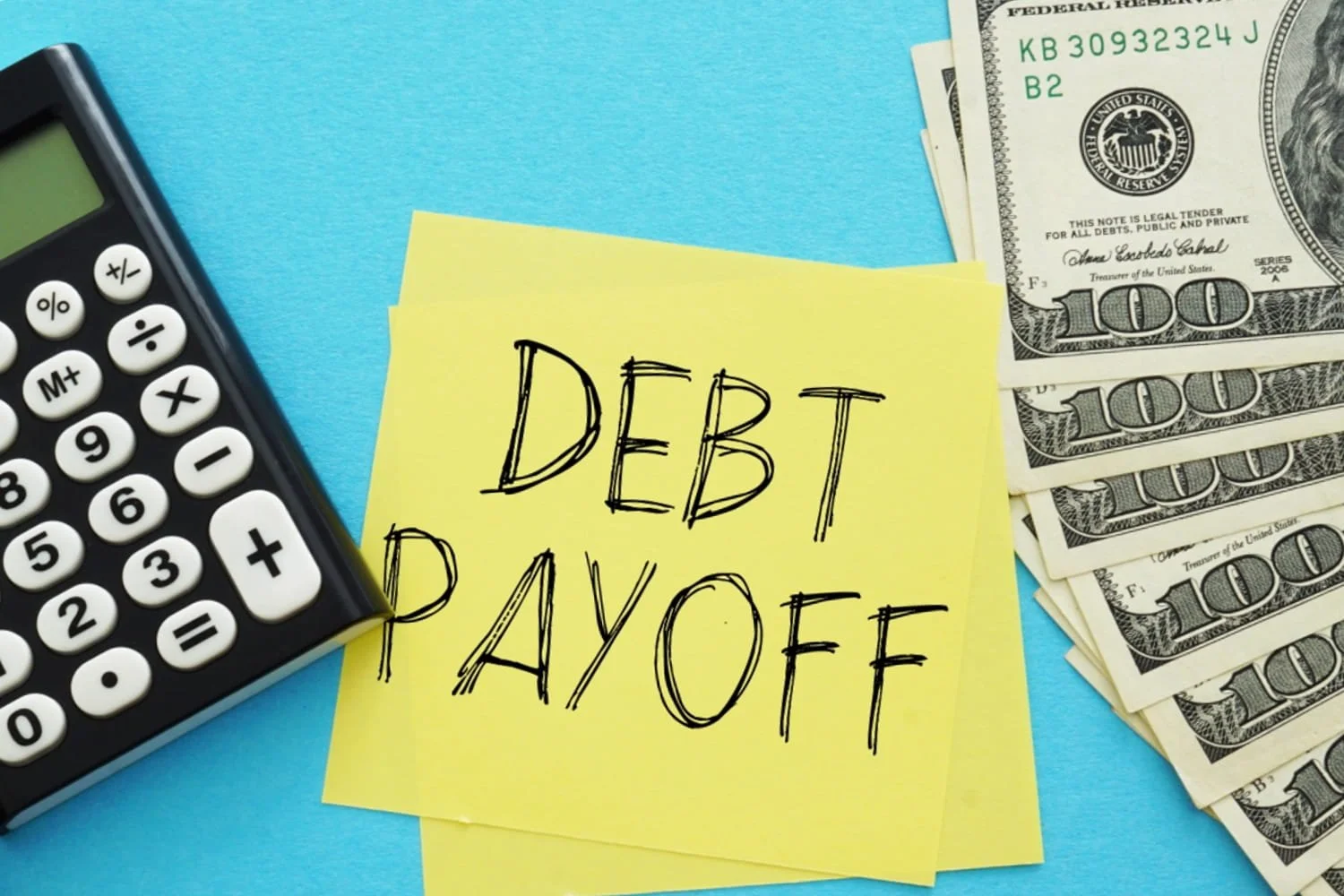 A yellow sticky note that says debt payoff with a calculator on the left and several U.S. one hundred dollar bills to the right.