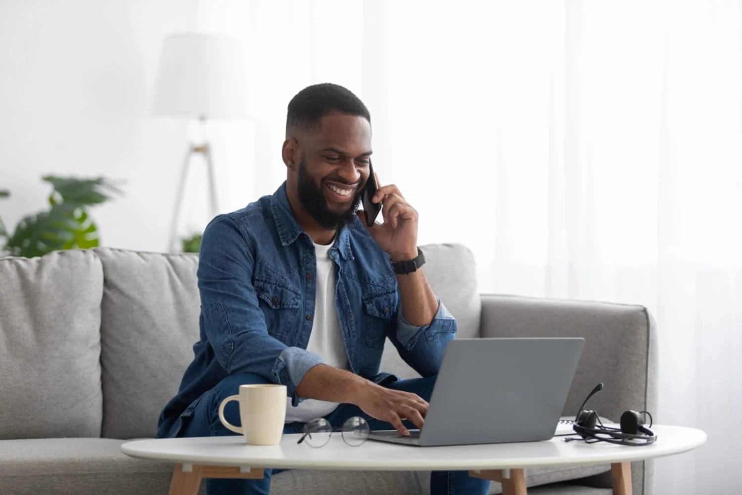 A man sitting on a sofa talking to someone on his cellular phone while using his laptop.