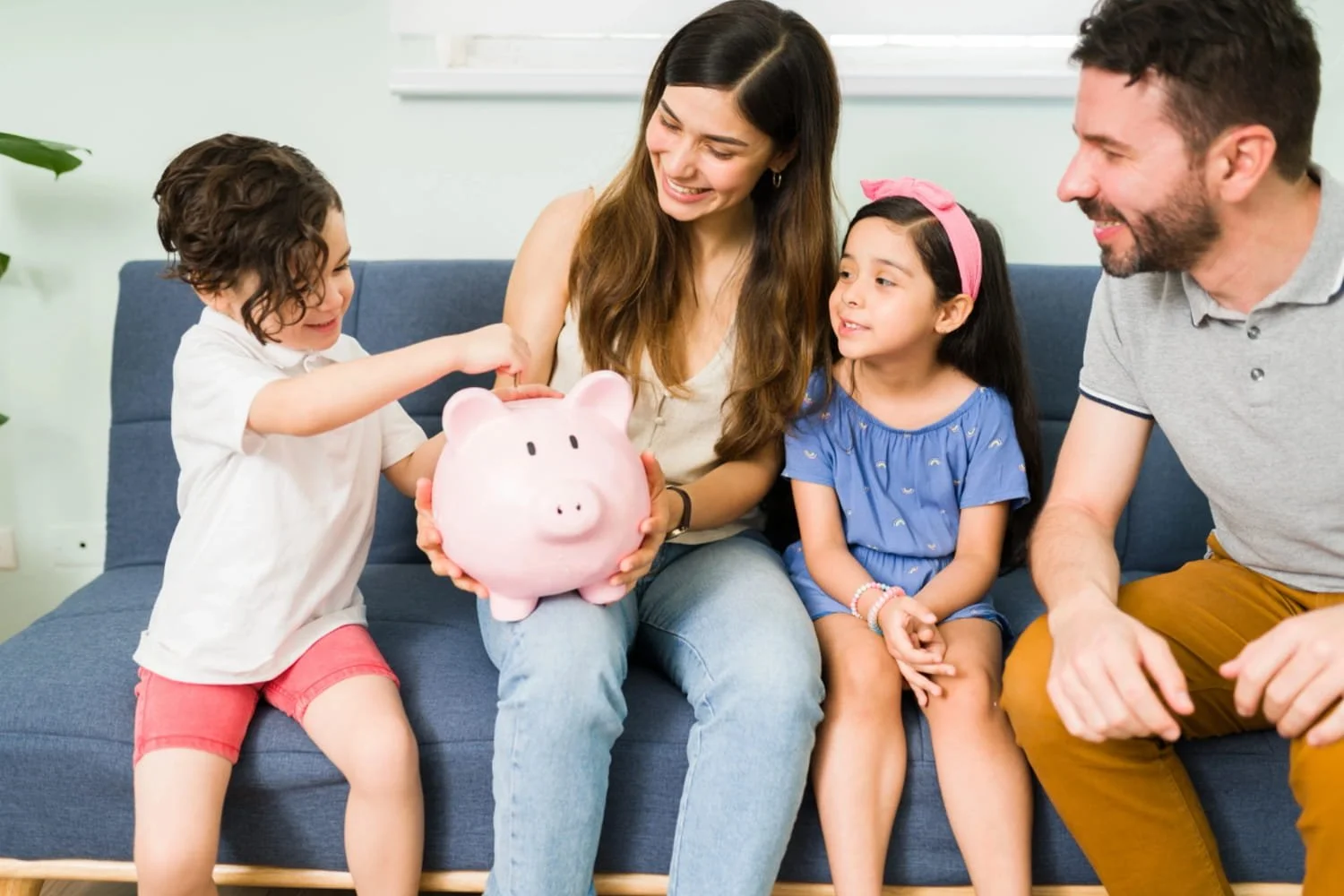 A mother and father sitting on a couch with their two young children and one child is placing a coin into a pink piggy bank that the mother is holding.