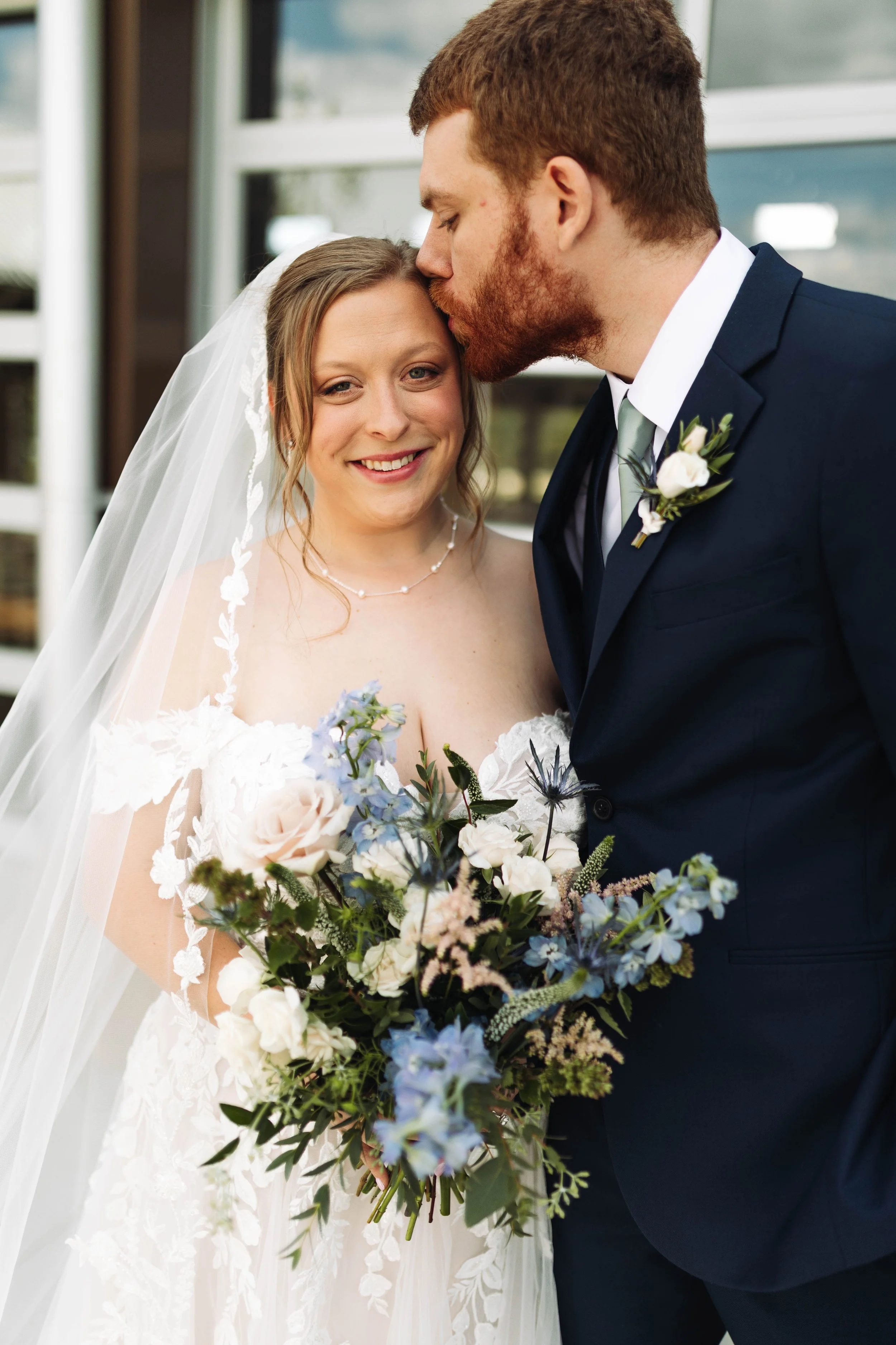 A bride with a white lace wedding dress and long veil, smiling and holding a bouquet of flowers, is kissed on the forehead by a groom in a dark blue suit with a white shirt and grey tie, with a boutonniere, in front of a glass building.