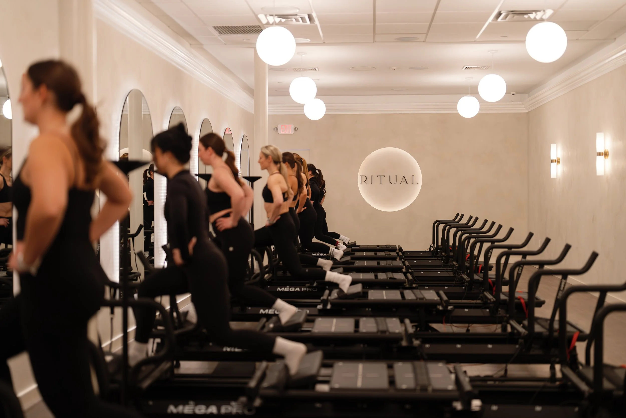 Group of women in black workout attire standing on Megaformers in a Lagree studio, ready for a class, with a sign saying 'RITUAL' on the wall.