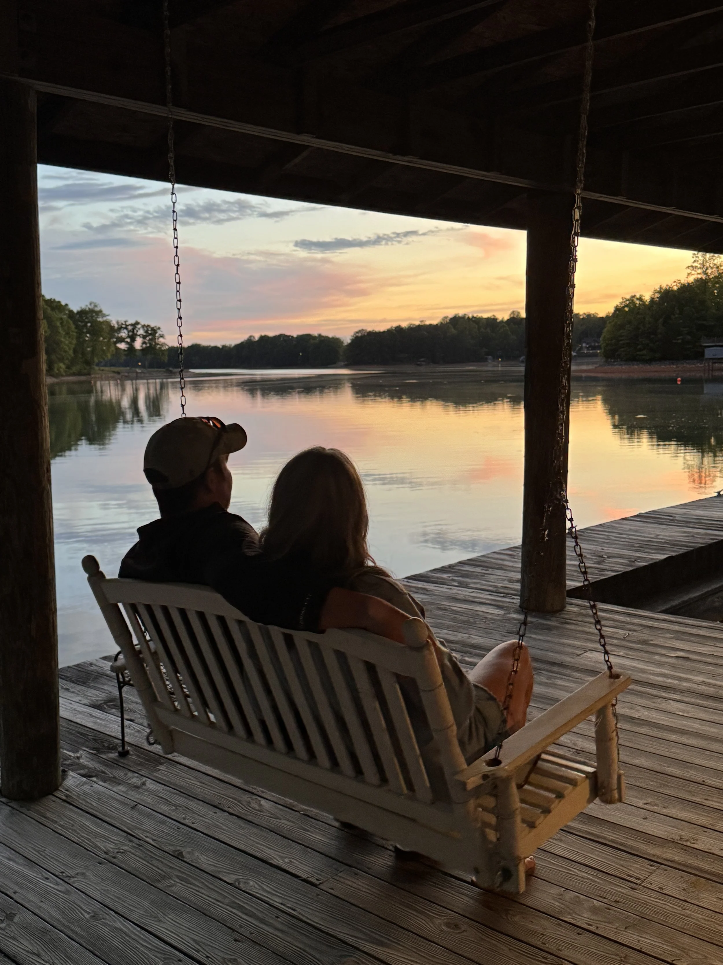 A couple sitting on a bench swing by a lake during sunset, with the sky showing hues of pink, orange, and blue, under a wooden pavilion.