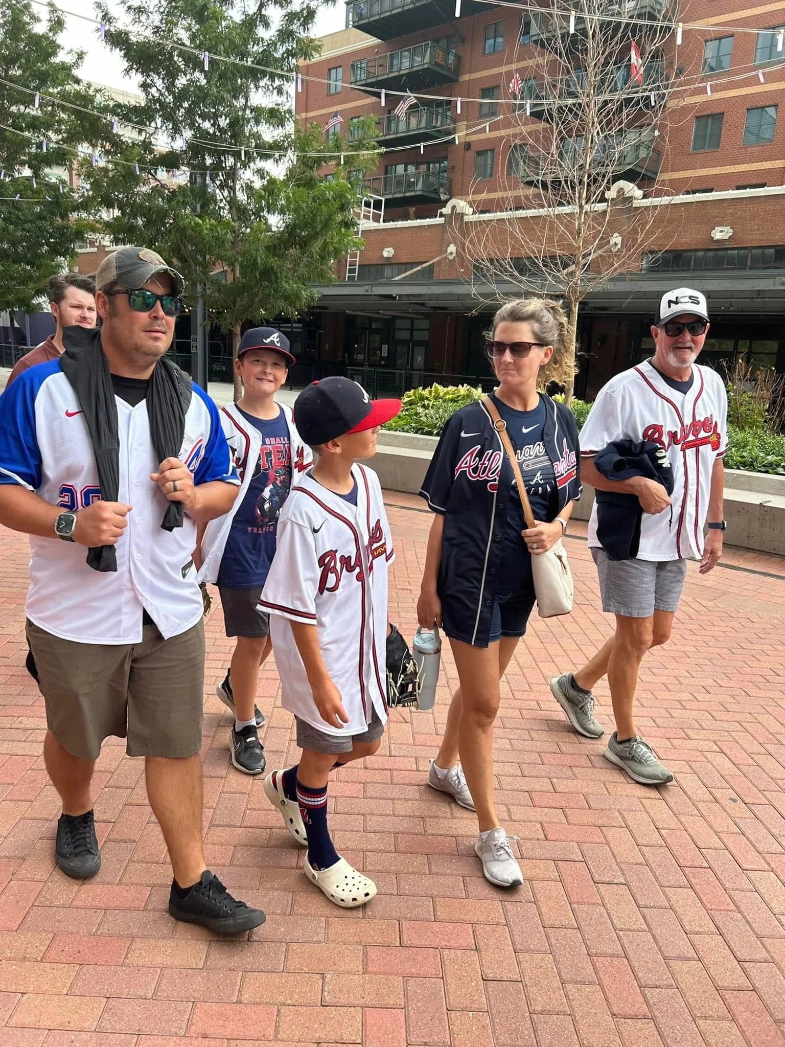 A group of six people, including children and adults, walking outdoors on a brick-paved path near a building with balconies. They are dressed in baseball jerseys, some with Atlanta Braves logos, and are wearing sunglasses. There are trees and flags hanging above them.