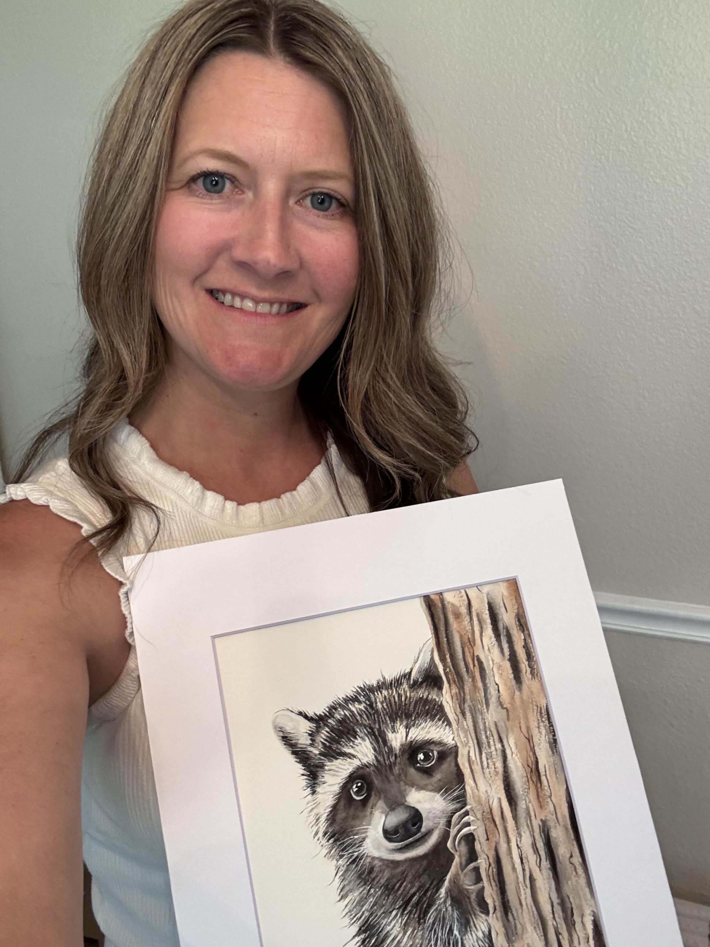 A woman with shoulder-length brown hair and blue eyes holding a poster with a detailed drawing of a raccoon peeking from behind a tree trunk.