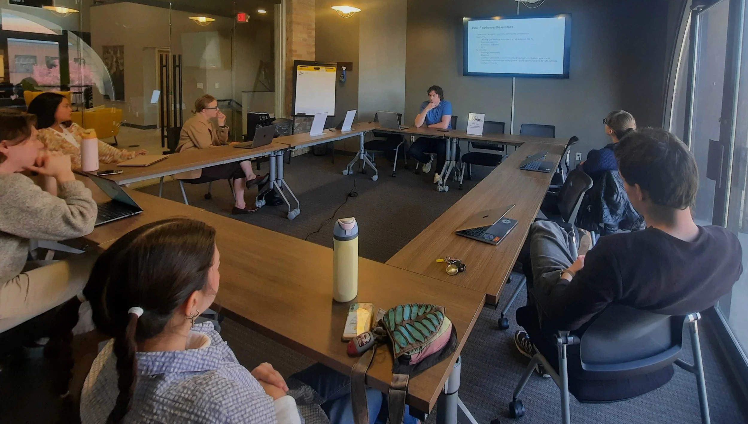 A group of people sit around a large square table. Fellow Ryan Kirsch sits at the front of the room, presenting. Ryan has dark curly hair and wears a blue shirt.