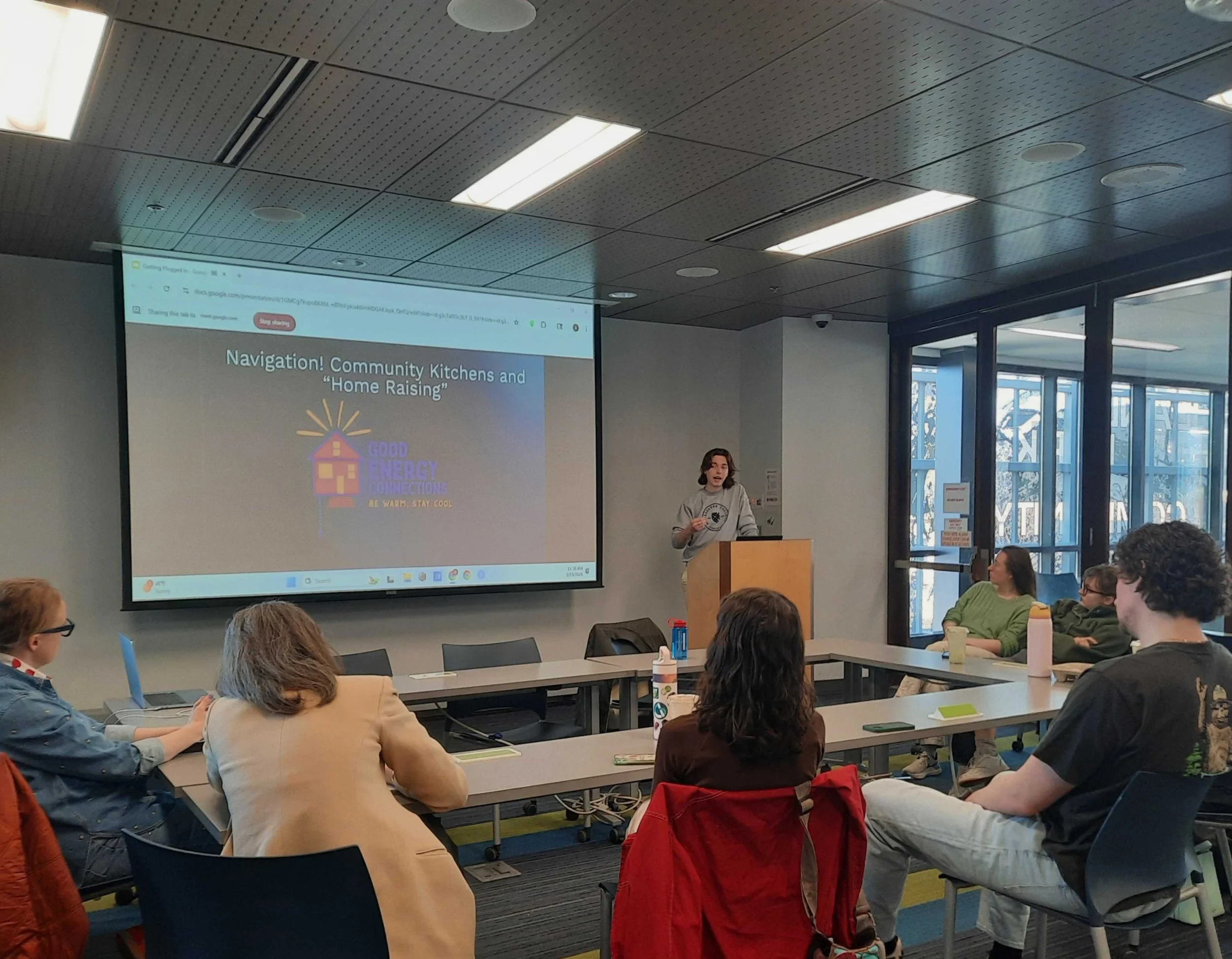 2025-26 Fellow Atticus Friendly speaks at a podium in a small meeting room. Bystanders look on.