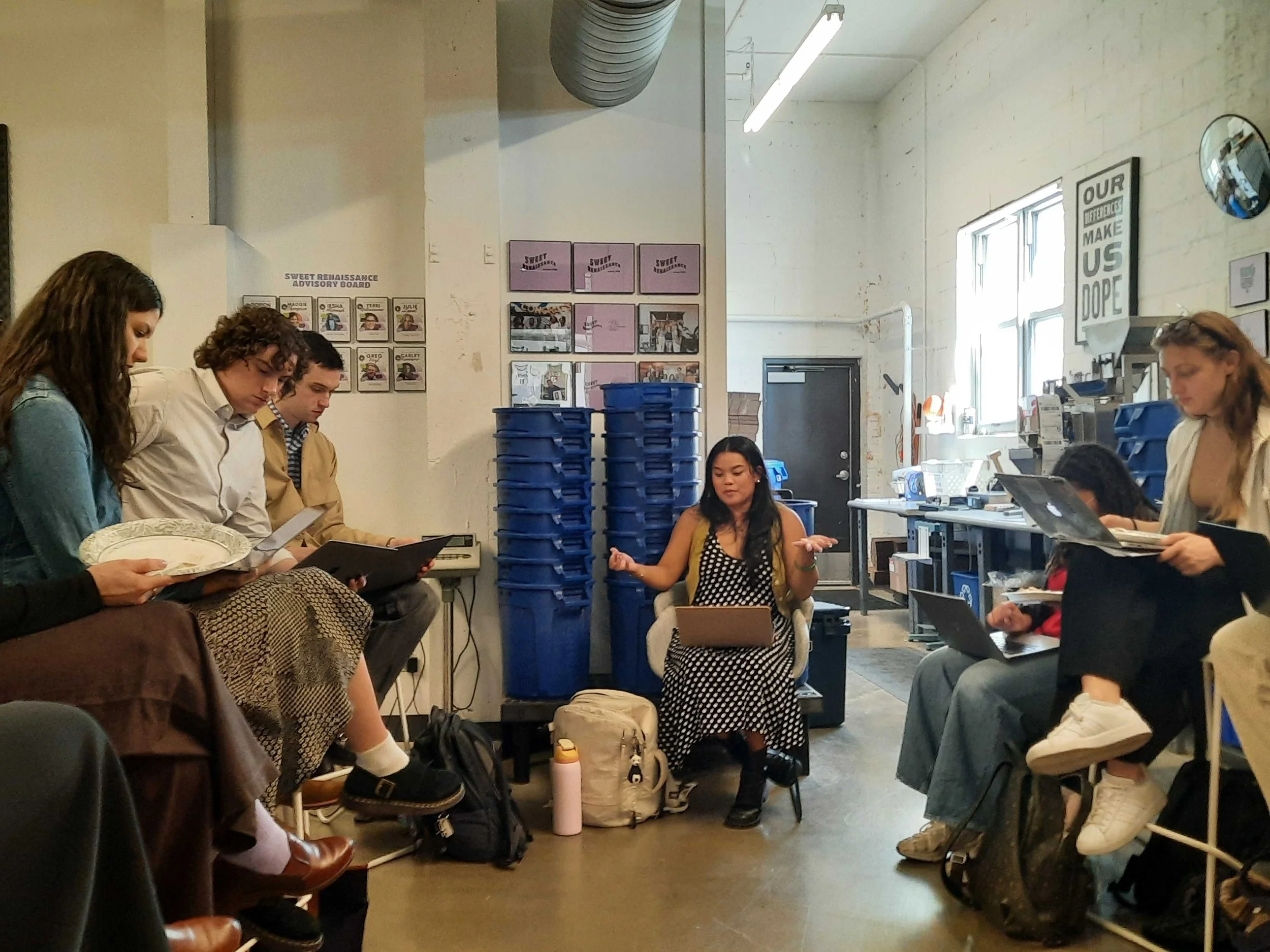 Fellow Ciboney Reglos sits in a chair with a laptop on her lap as she speaks to a group of people. She has long dark hair and wears a black and white polka dot dress.