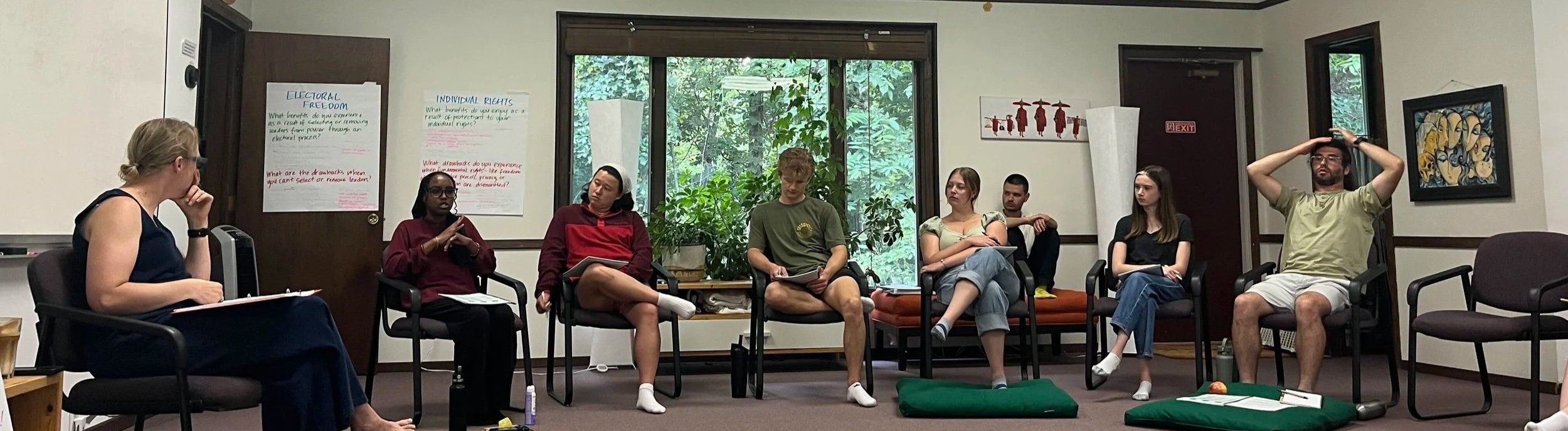 A group of fellows sits in a half circle and listens to one of their peers. Jill Carey sits at the front.
