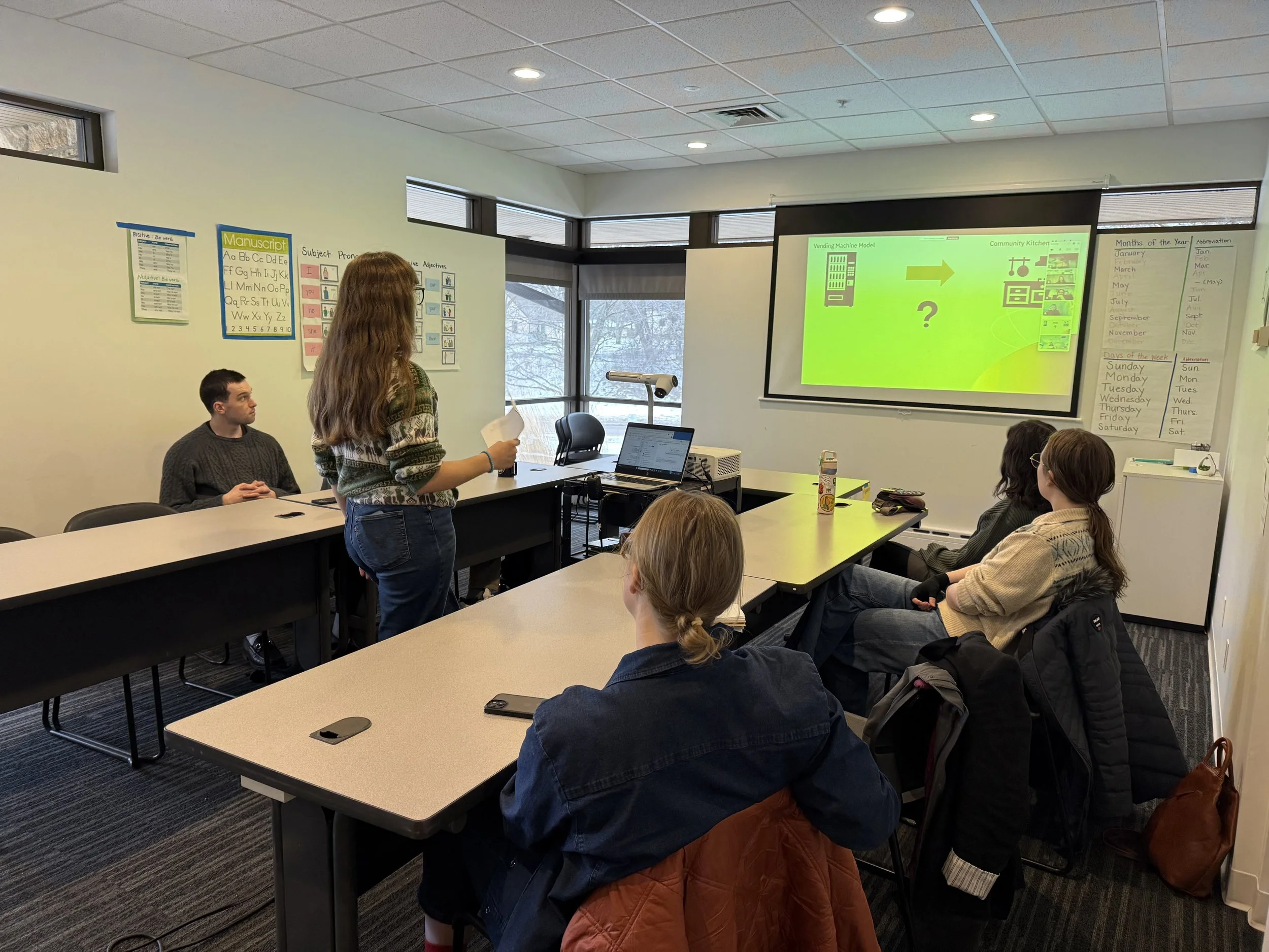 Madison Studer presents to the Civic Bridgers Fellowship cohort. She stands in front of a projector screen as multiple people look on. Snow is visible through the window.