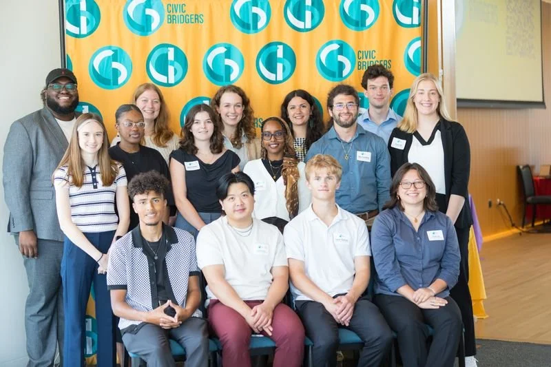 A group of 15 young adults arranged in three rows smiles at the camera. There is a golden yellow backdrop behind them patterned with Civic Bridgers logos.