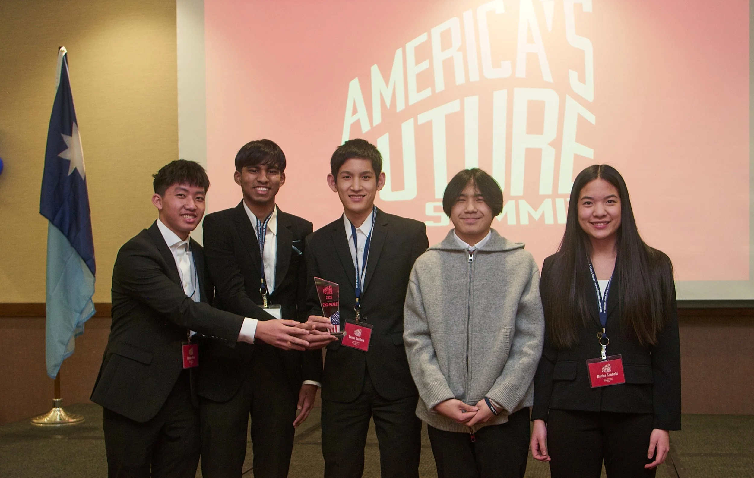 A group of 5 young people smile at the camera, holding a glass trophy. The are all wearing dark suits.