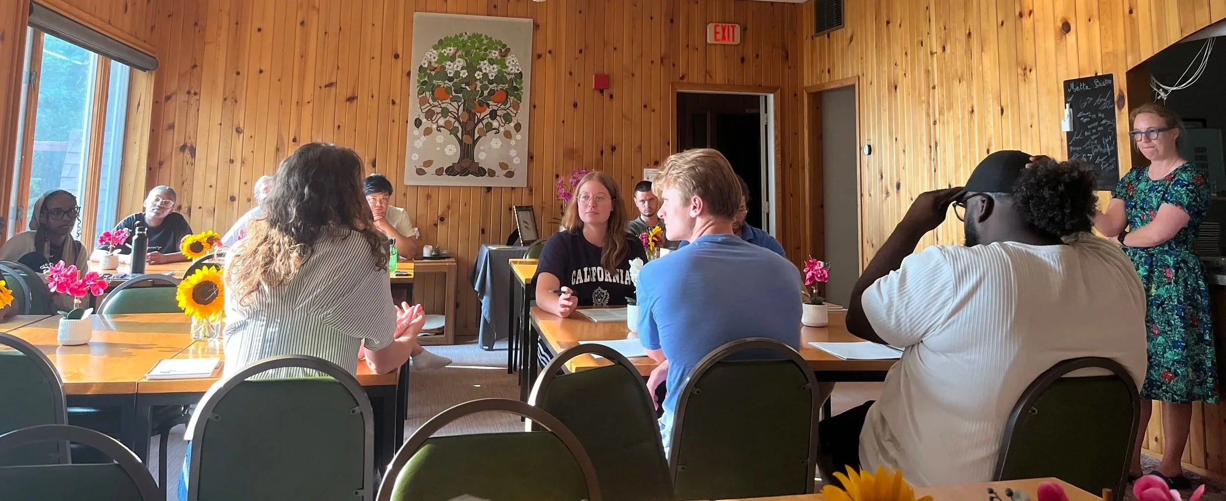 Fellows sit around small wooden tables and listen to one of their peers.