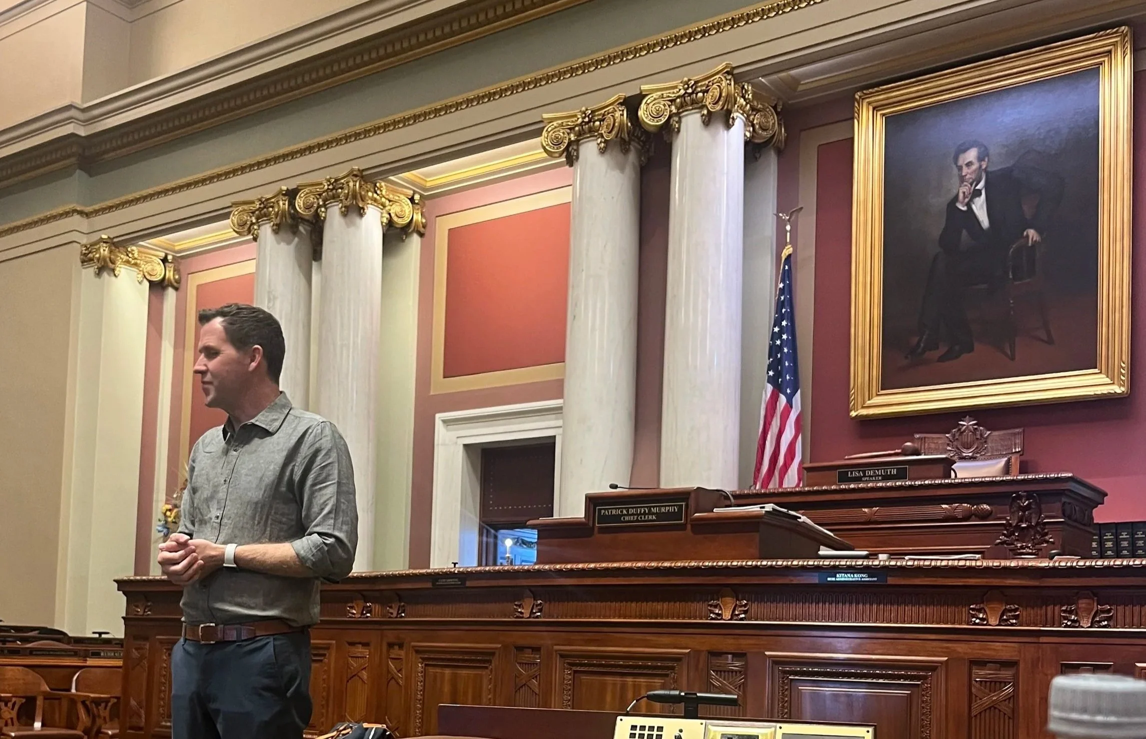 Minnesota House Rep. Jamie Long speaks on the House floor in front of the Speaker's podium. A portrait of Abraham Lincoln hangs on the wall.