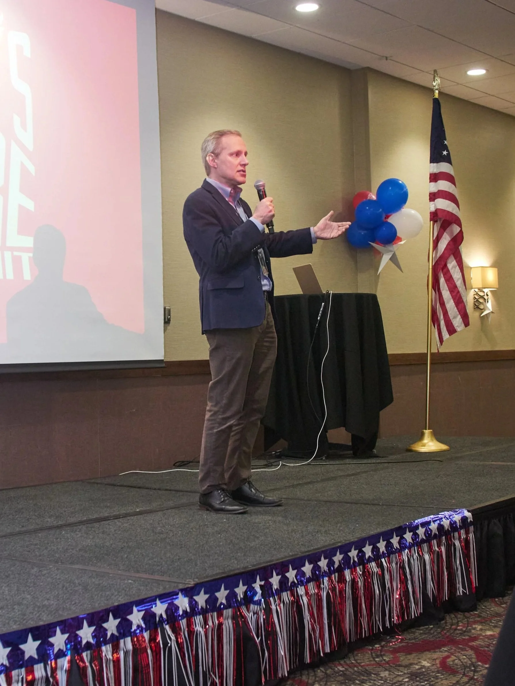 Steve Simon speaks into a microphone while standing on a stage. He wears a navy jack, blue shirt, and brown pant.