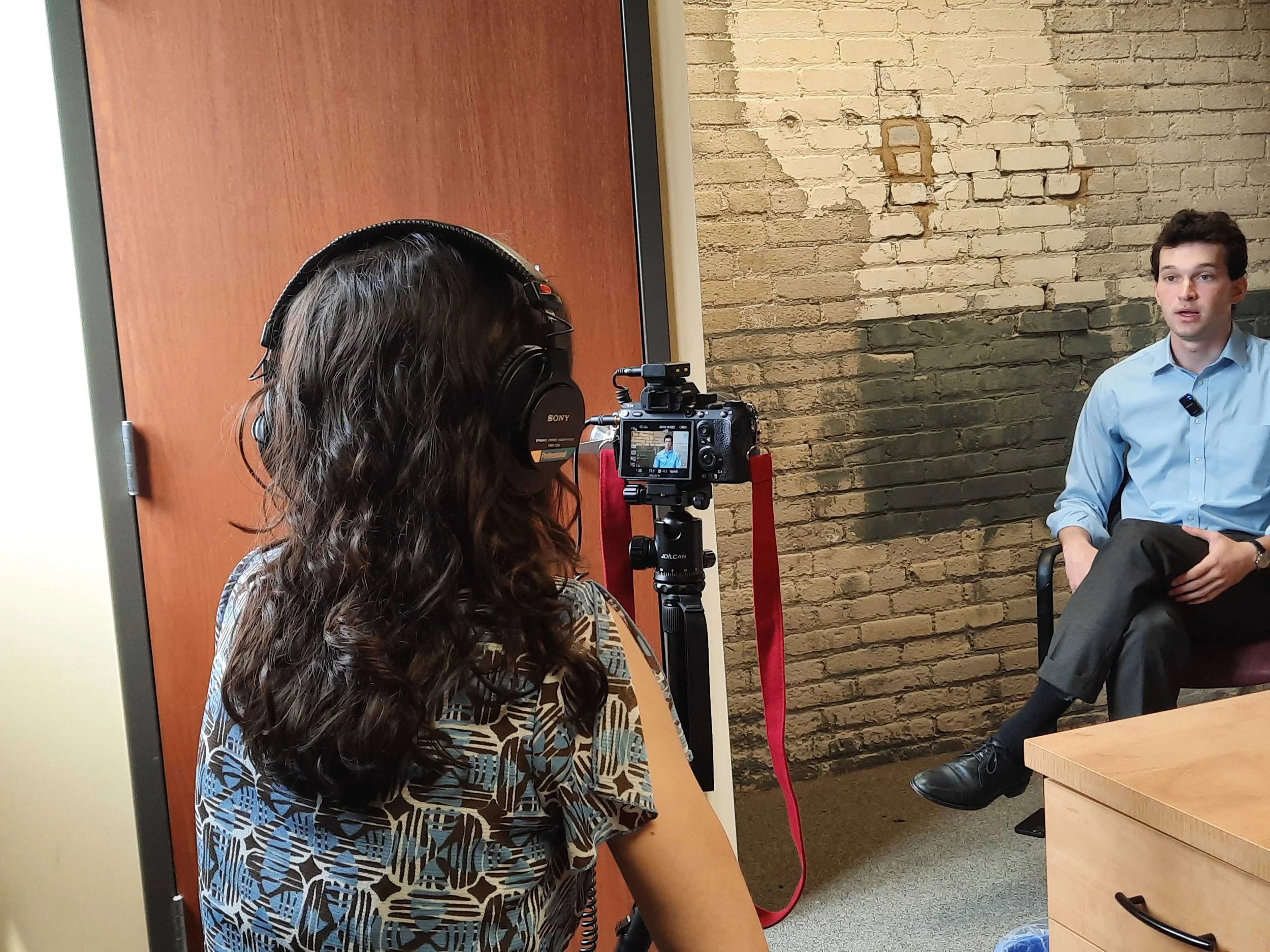 Fellow Taylor Canas sits behind a camera with black headphones on. In the photo you can see her subject as he speaks to the camera. Taylor has long dark hair and wears a blue and brown patterned shirt.