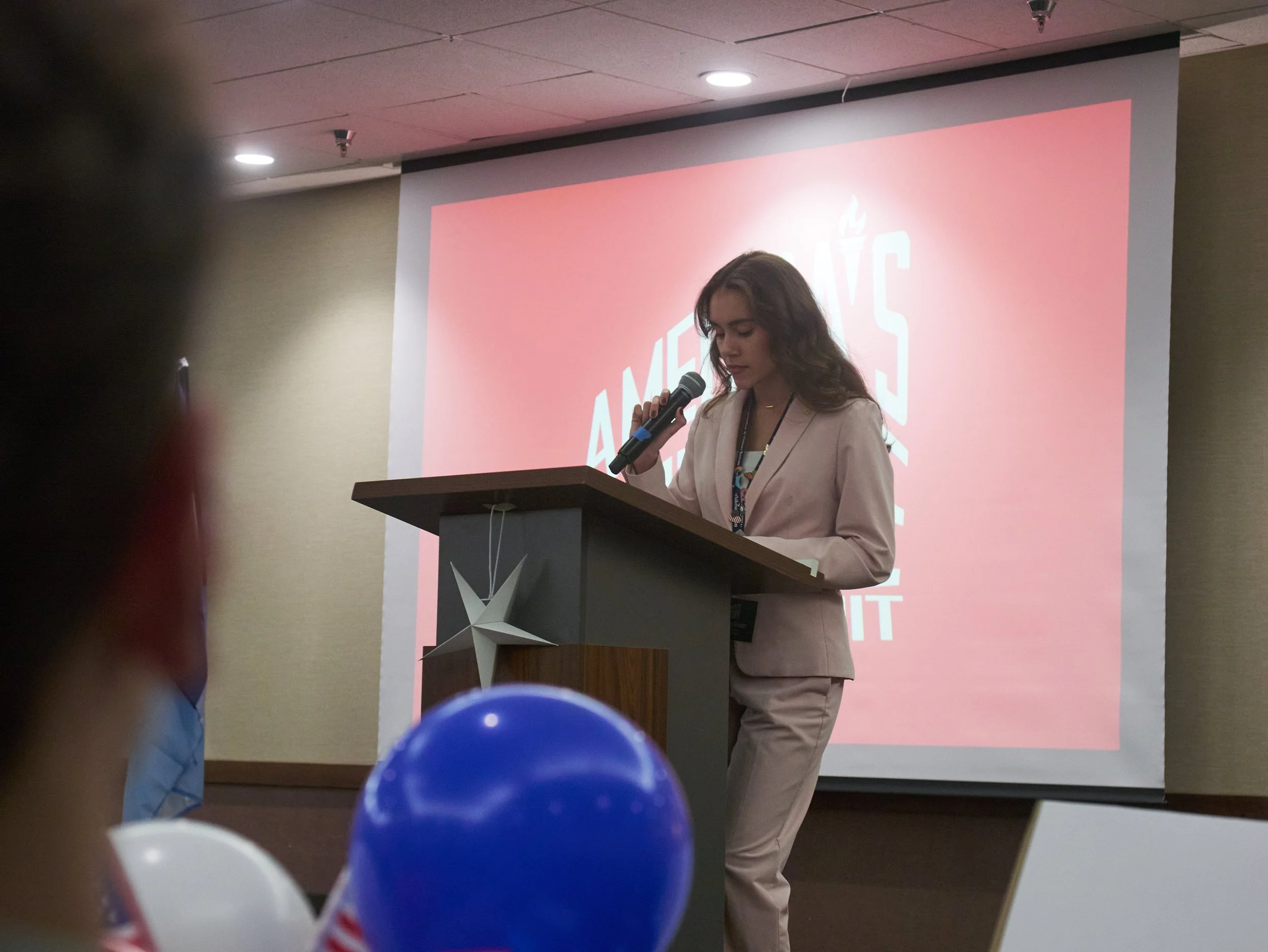 Maitreya Reeder stands at a podium with a microphone in hand. She wears a pink suit and has long brown hair.