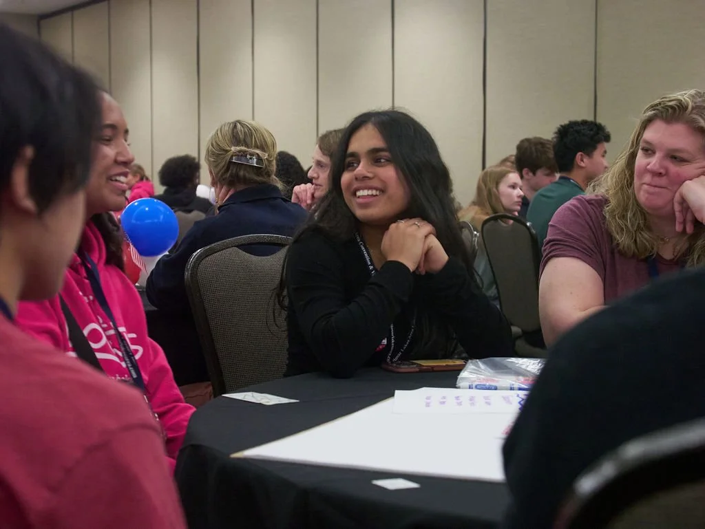 A young woman looks to her right at another student. They sit at a table with various papers on top of it. Their is an active conversation happening.