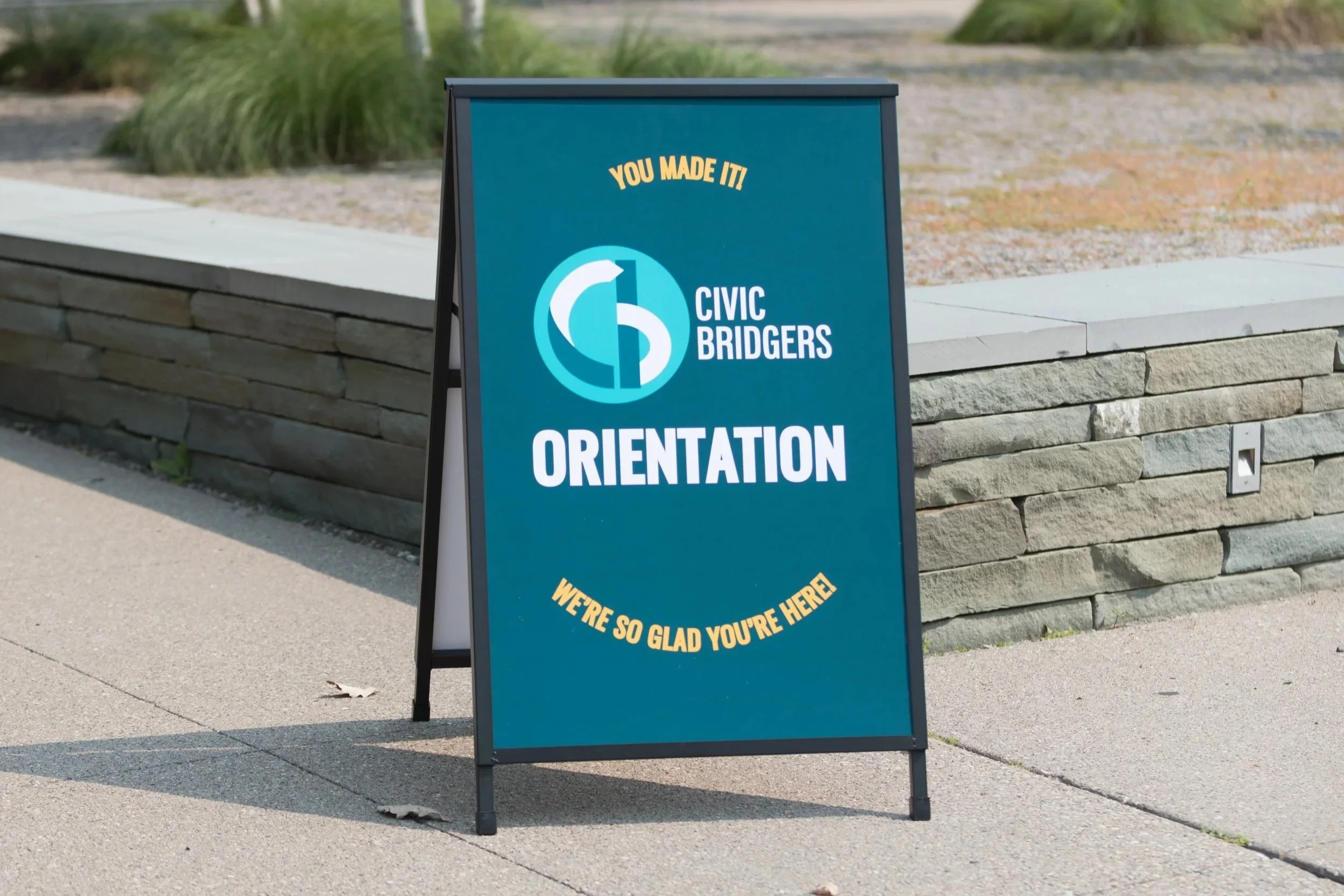 A teal sandwich board style sign on the sidewalk reads "You made it! Civic Bridgers Orientation. We're so glad you're here!"