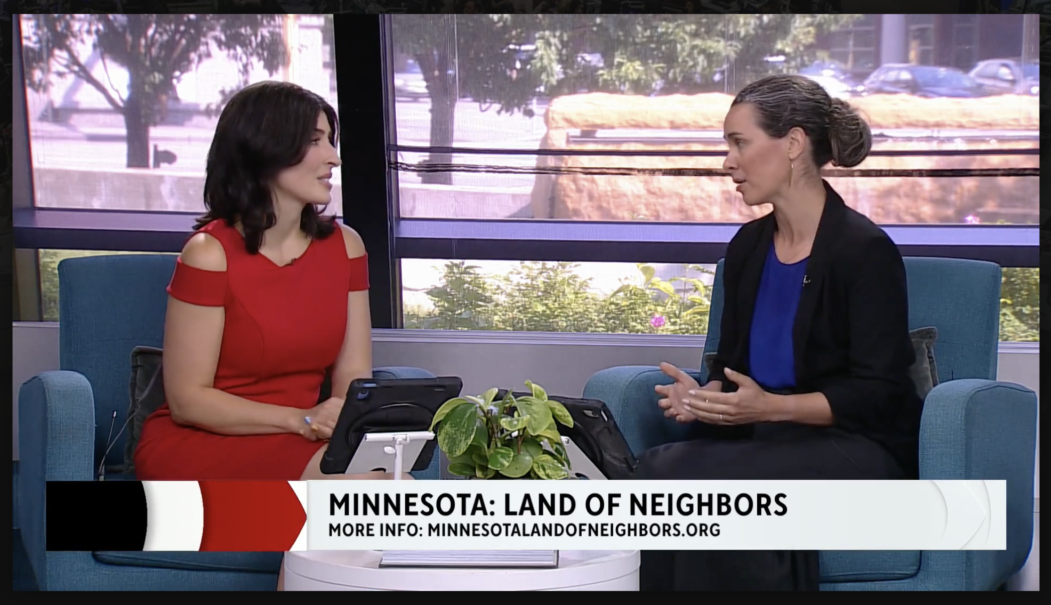 Libby Stegger sits in a blue couch. Shhe has dark hair pulled back and wears a blue and black shirt. She sits opposite a WCCO host, wearing a red dress and sitting in a blue chair.