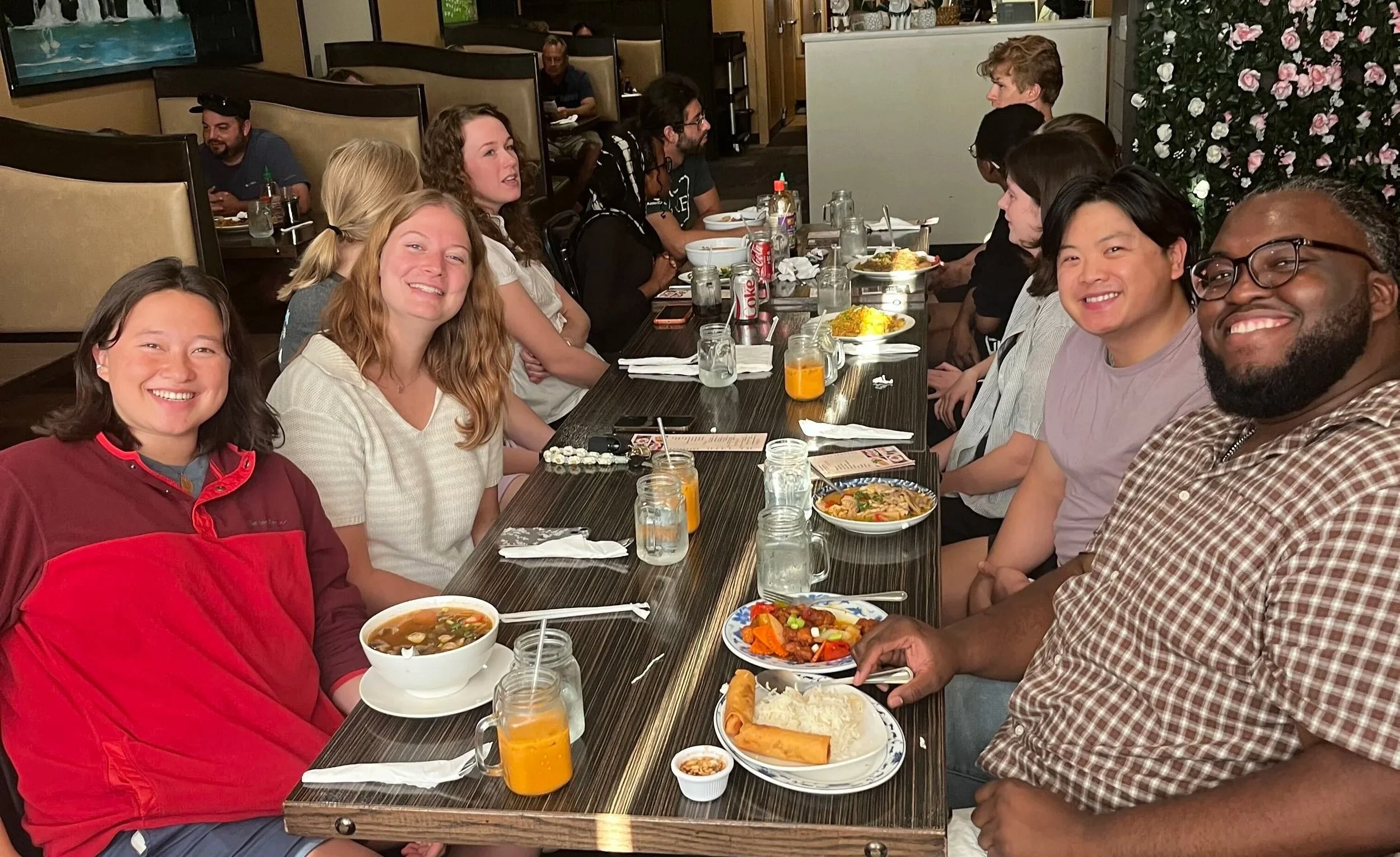 Four Fellows, including Dez Harris, smile at the camera. They are sitting around a table getting ready to eat dinner. Other Fellows talk in the background.