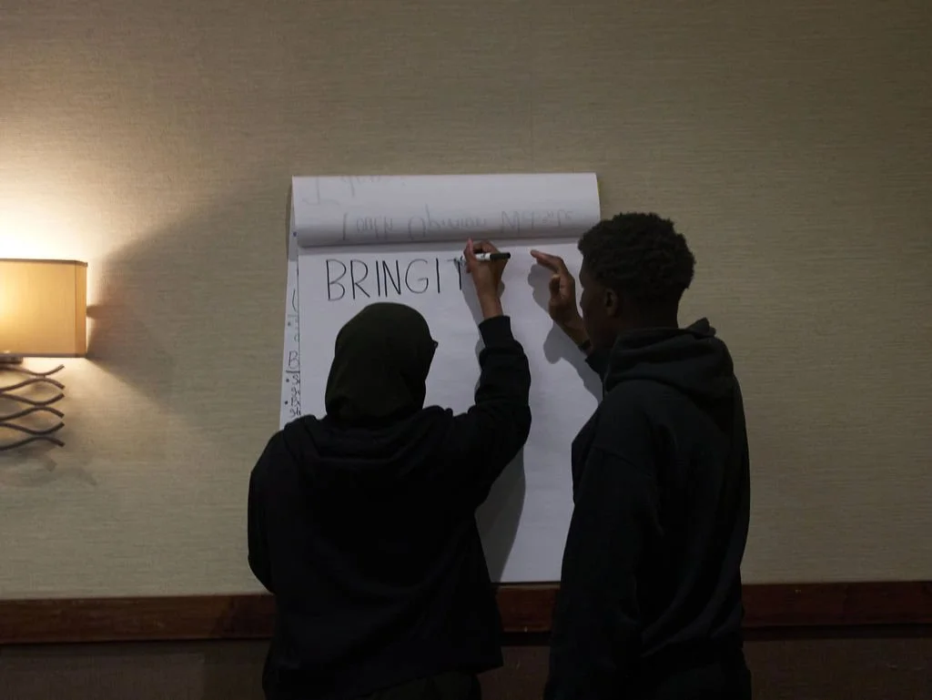 2 young people write on a piece of large poster paper. Their backs are to the camera. The words visible on the poster are "BRINGIT"