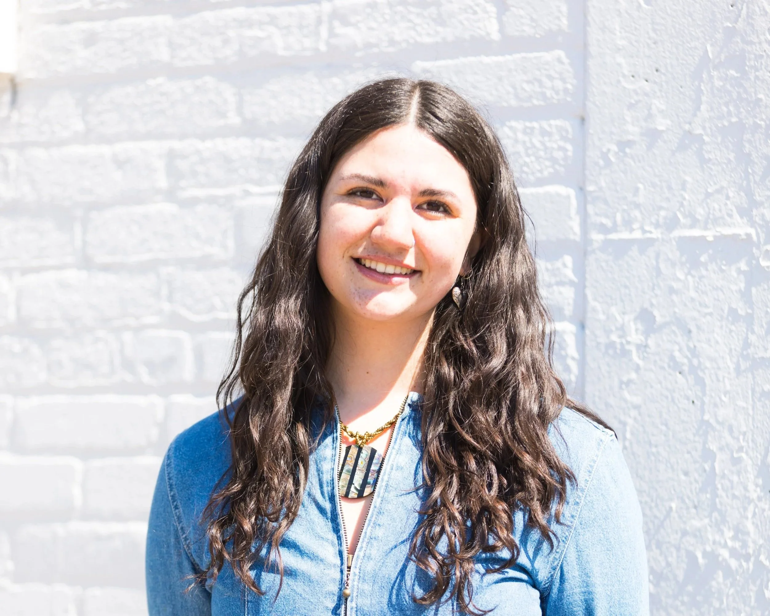A headshot of Senior Fellow, Angelina, in a denim top with a white brick wall as the backdrop.