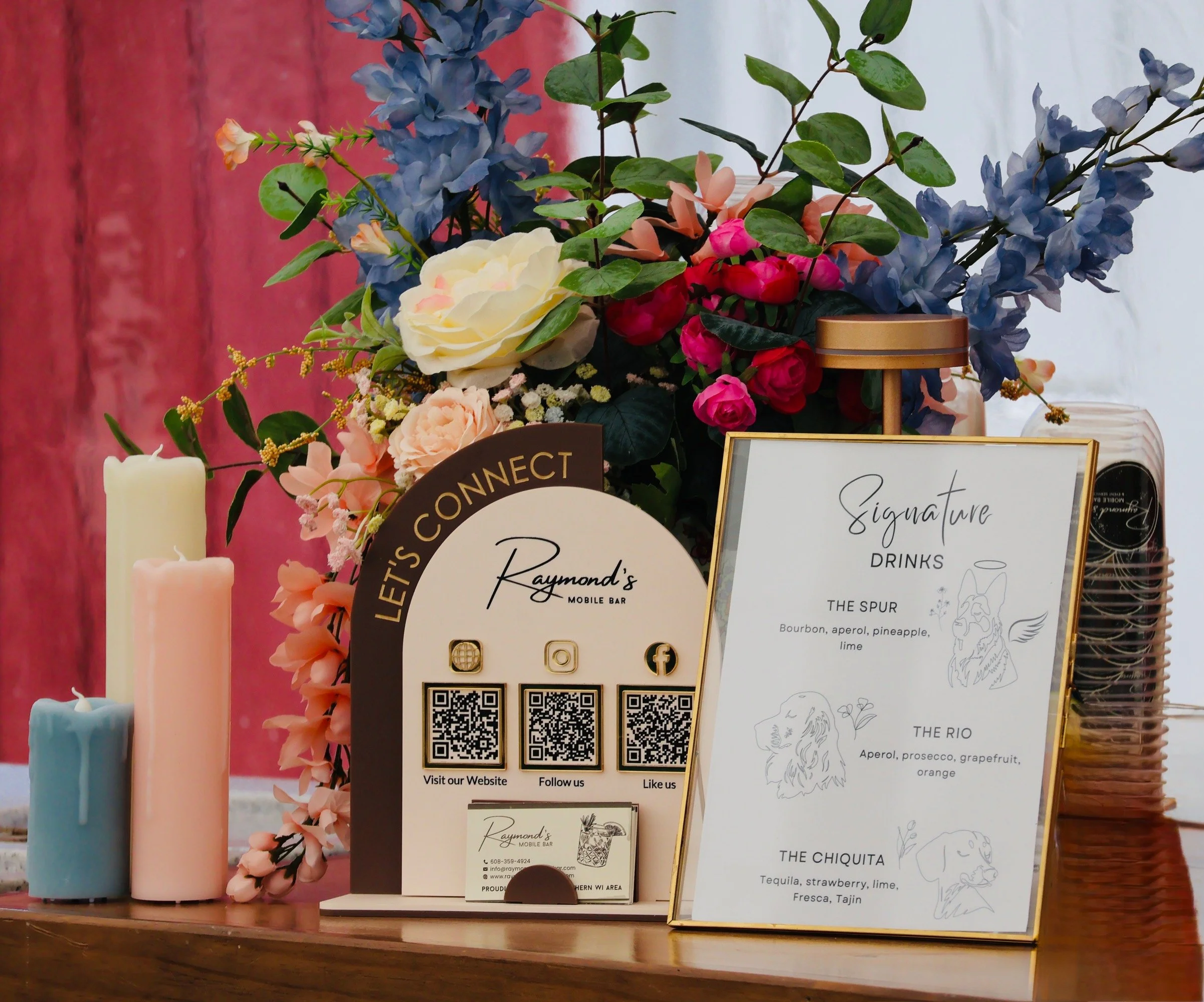 A floral arrangement with blue, pink, cream, and peach-colored flowers, displayed on a table with a pink wooden background. There are candles nearby and informational signs with QR codes and handwritten menus for drinks, including signature drinks, t