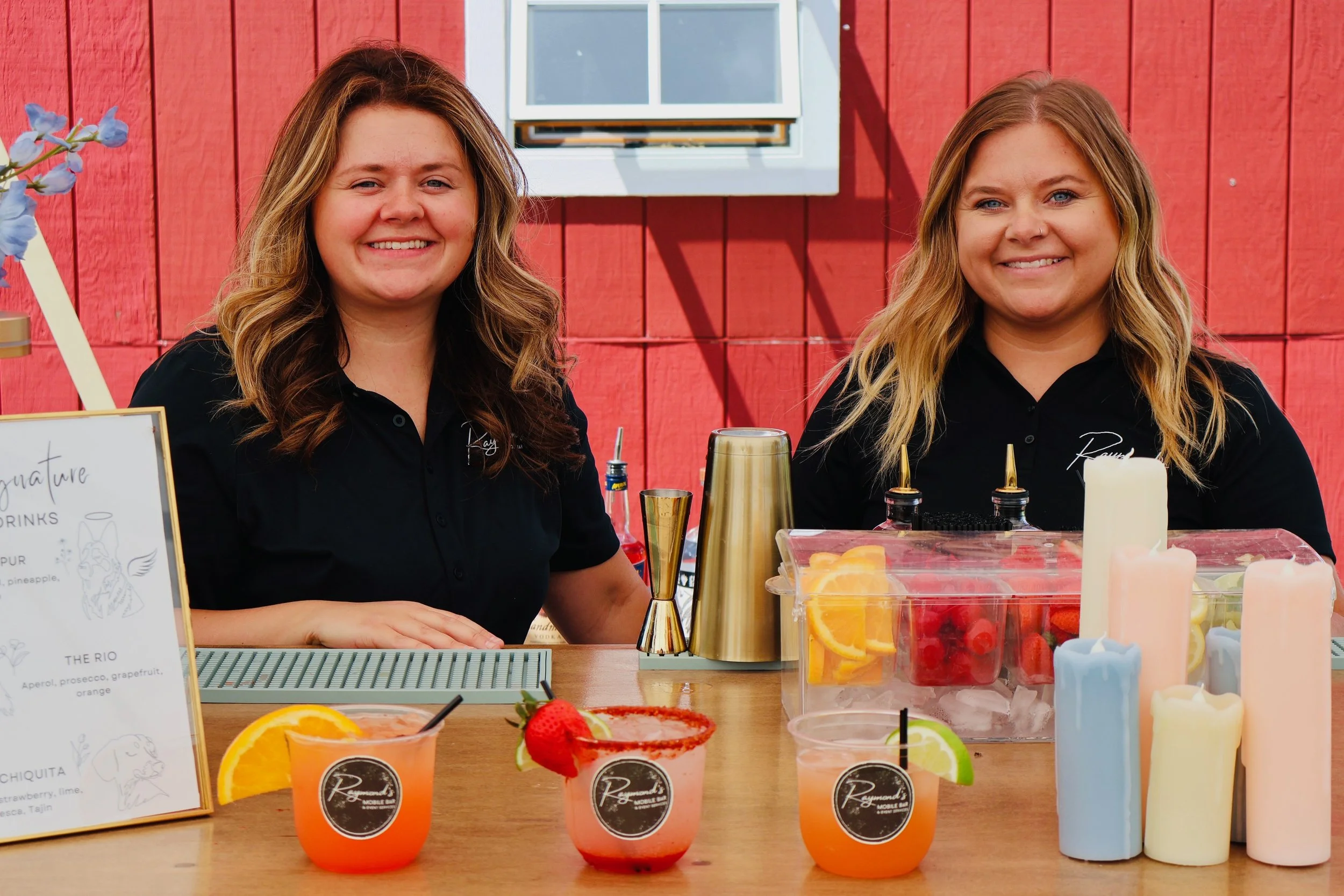 Two smiling women behind a bar with colorful cocktail drinks, fresh fruit slices, and whipped cream on the table, against a red wooden wall.