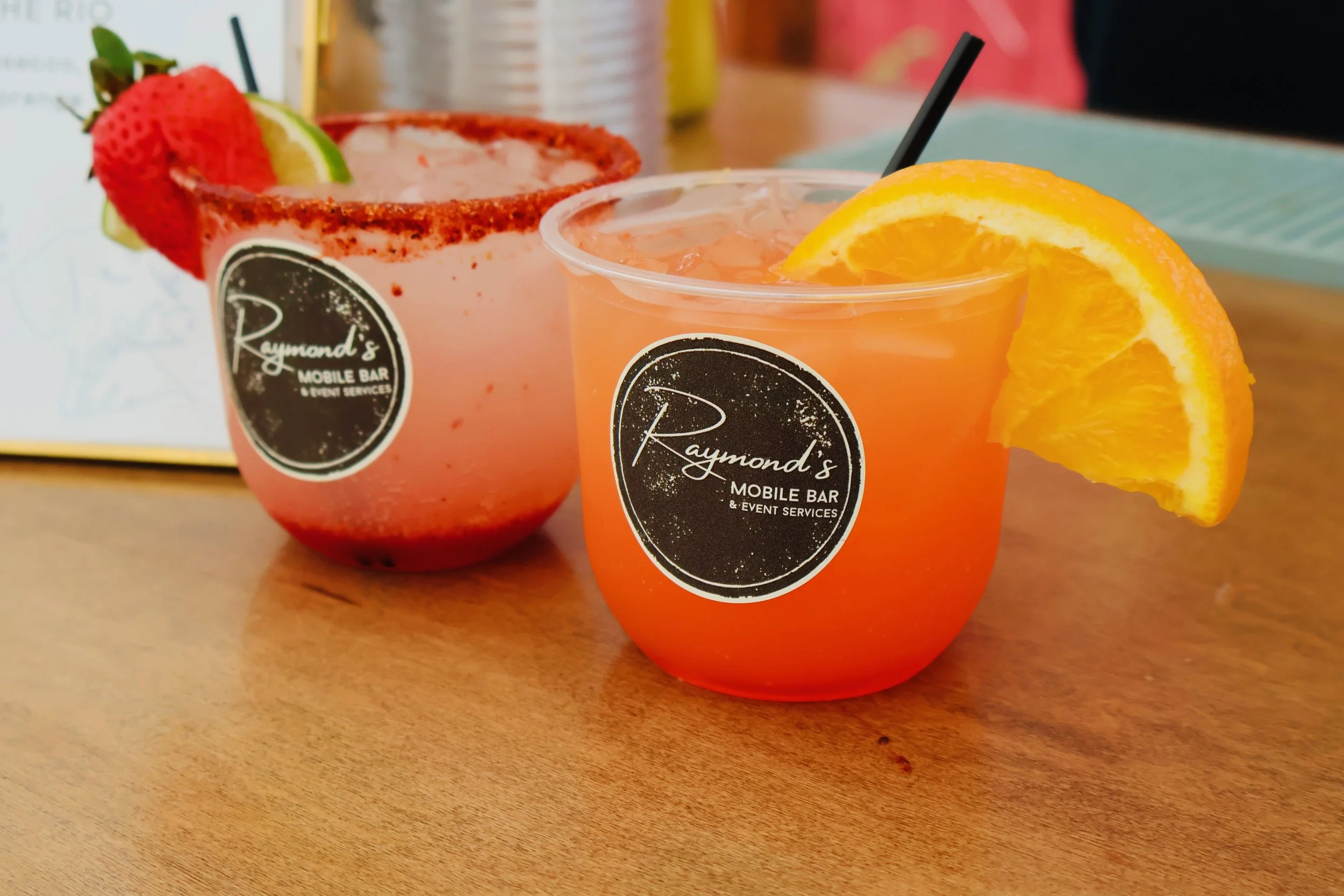 Two drinks in clear cups with black straws on a wooden table, garnished with orange slices on one and strawberries and lime on the other, with a logo reading 'Raymond's Mobile Bar & Event Services'.