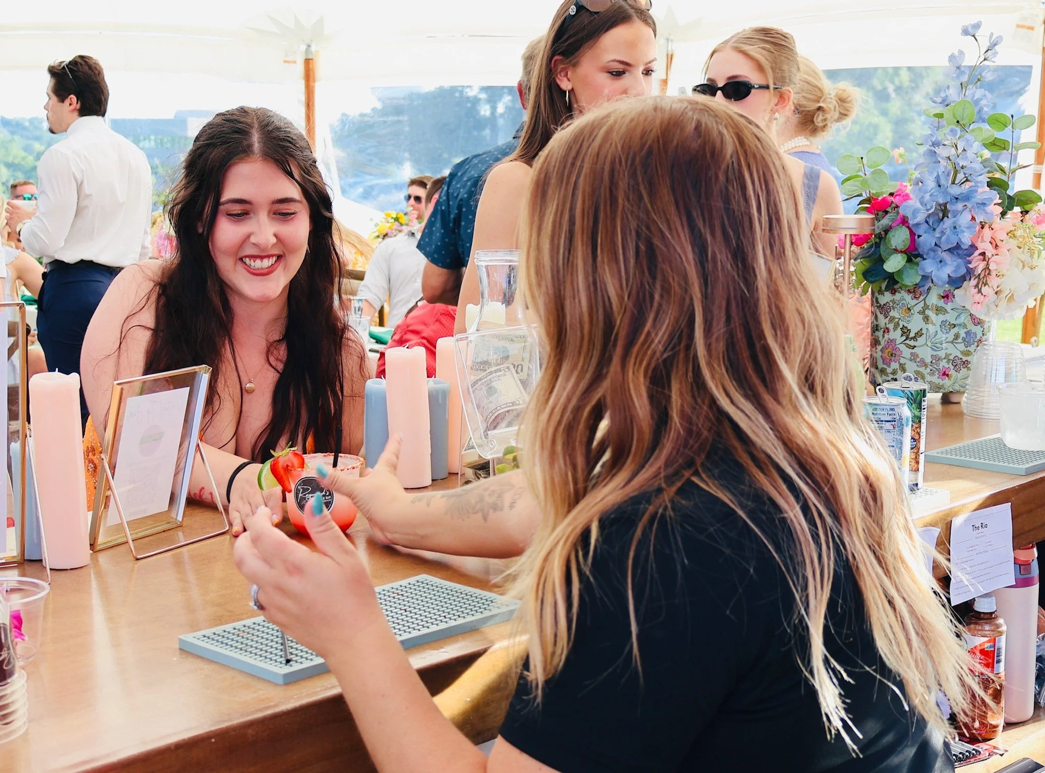 People socializing and enjoying drinks at an outdoor event or party under a tent, with floral decorations and a scenic landscape in the background.