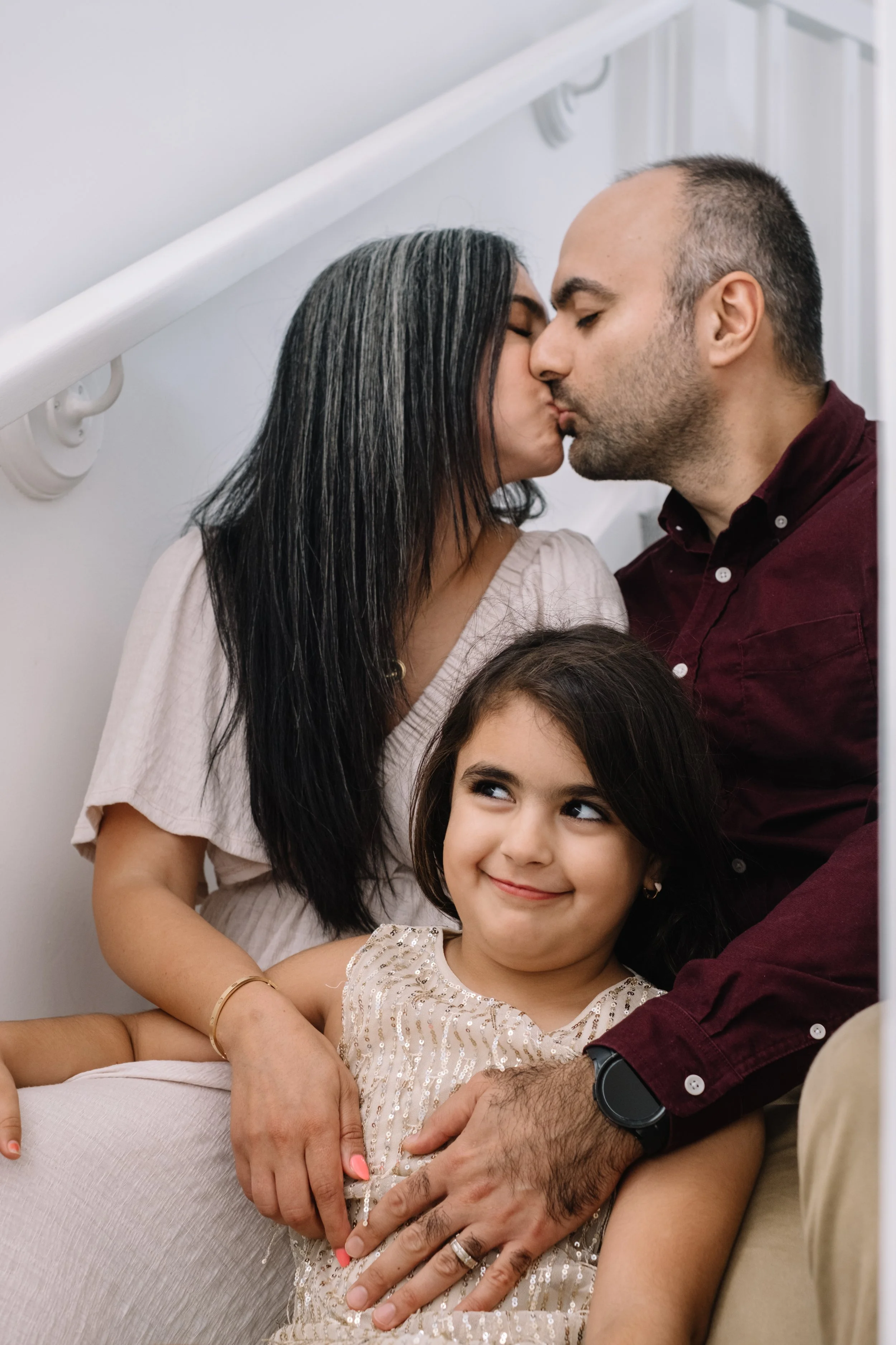 A family of three: a woman, a man, and a young girl. The woman and man are kissing, while the girl, sitting on her mother's lap, is smiling and looking to the side.