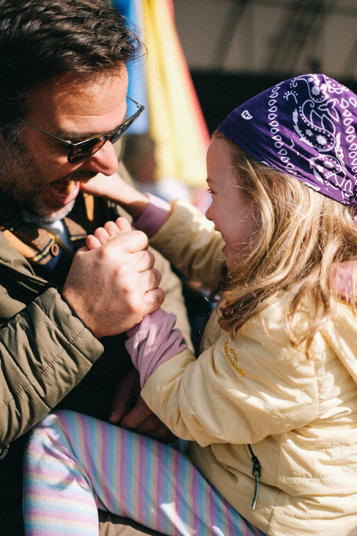 A man and a young girl smiling and holding hands, enjoying a moment outdoors during daytime, with colorful flags in the background.