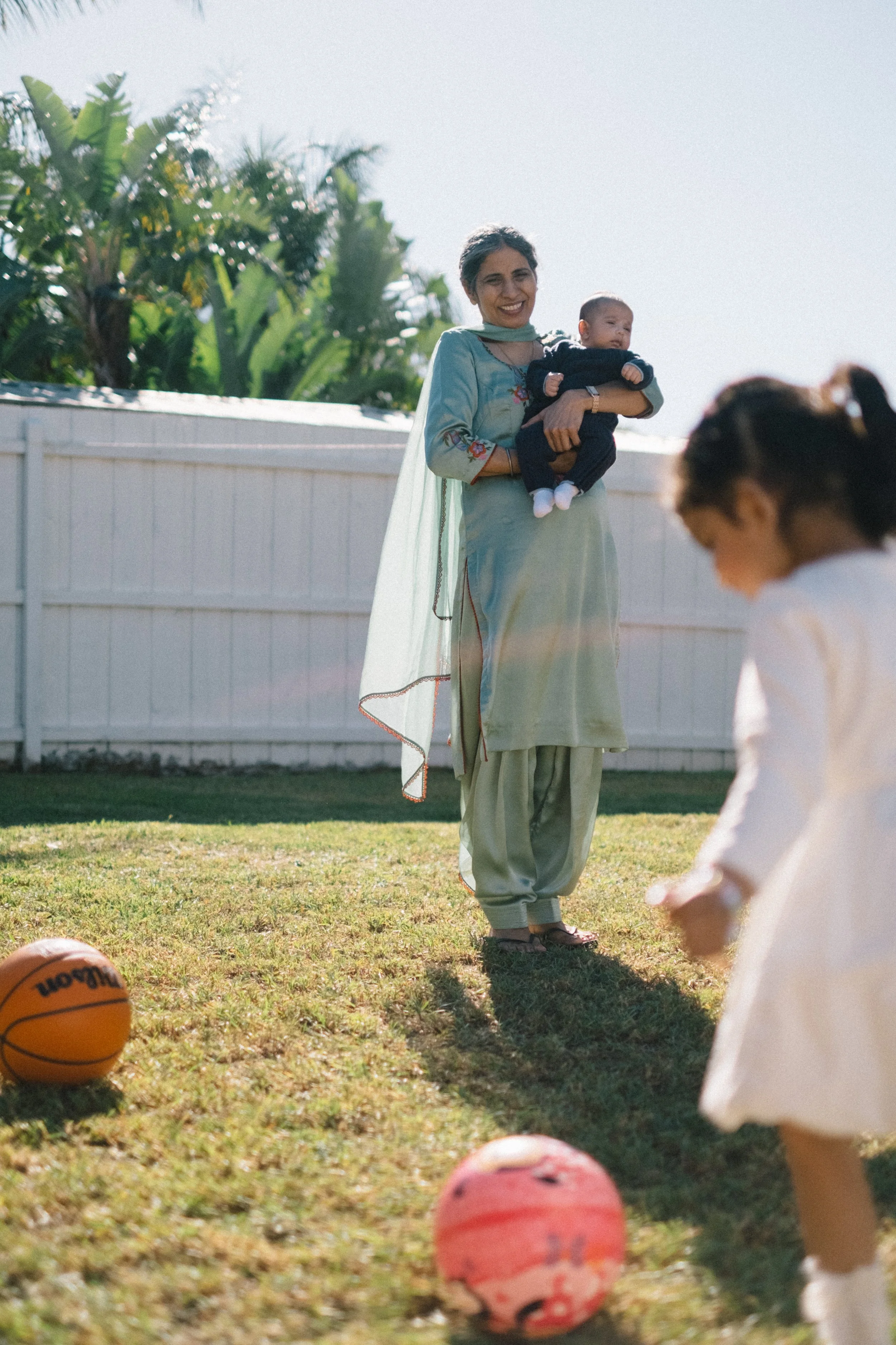 A woman in traditional South Asian attire holding a baby outdoors during daytime, with children playing nearby and basketballs on the grass.