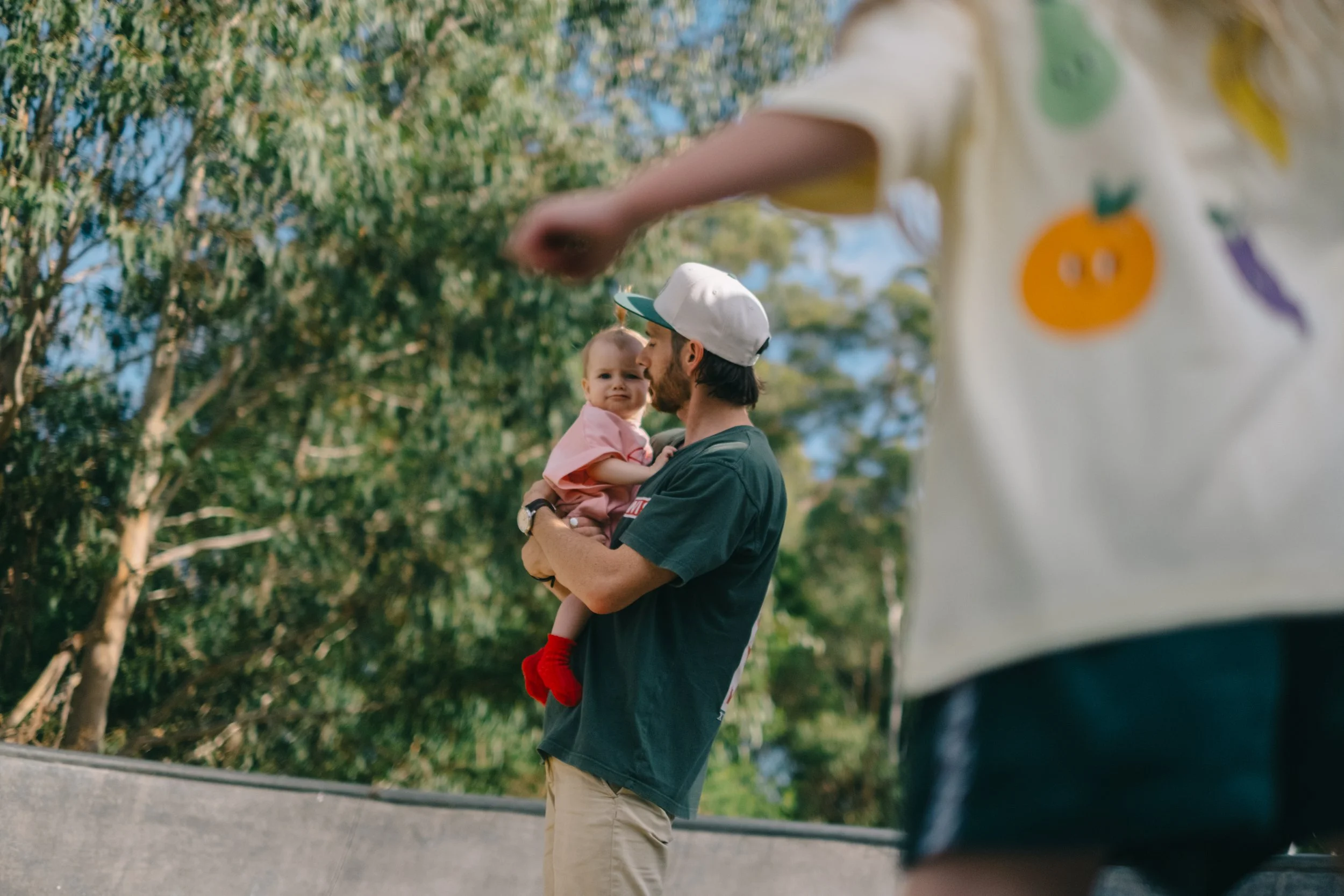 A man is holding a young girl in a pink shirt and red socks outdoors with trees in the background. Another person wearing a white shirt with colorful fruit designs is partially visible in the foreground.