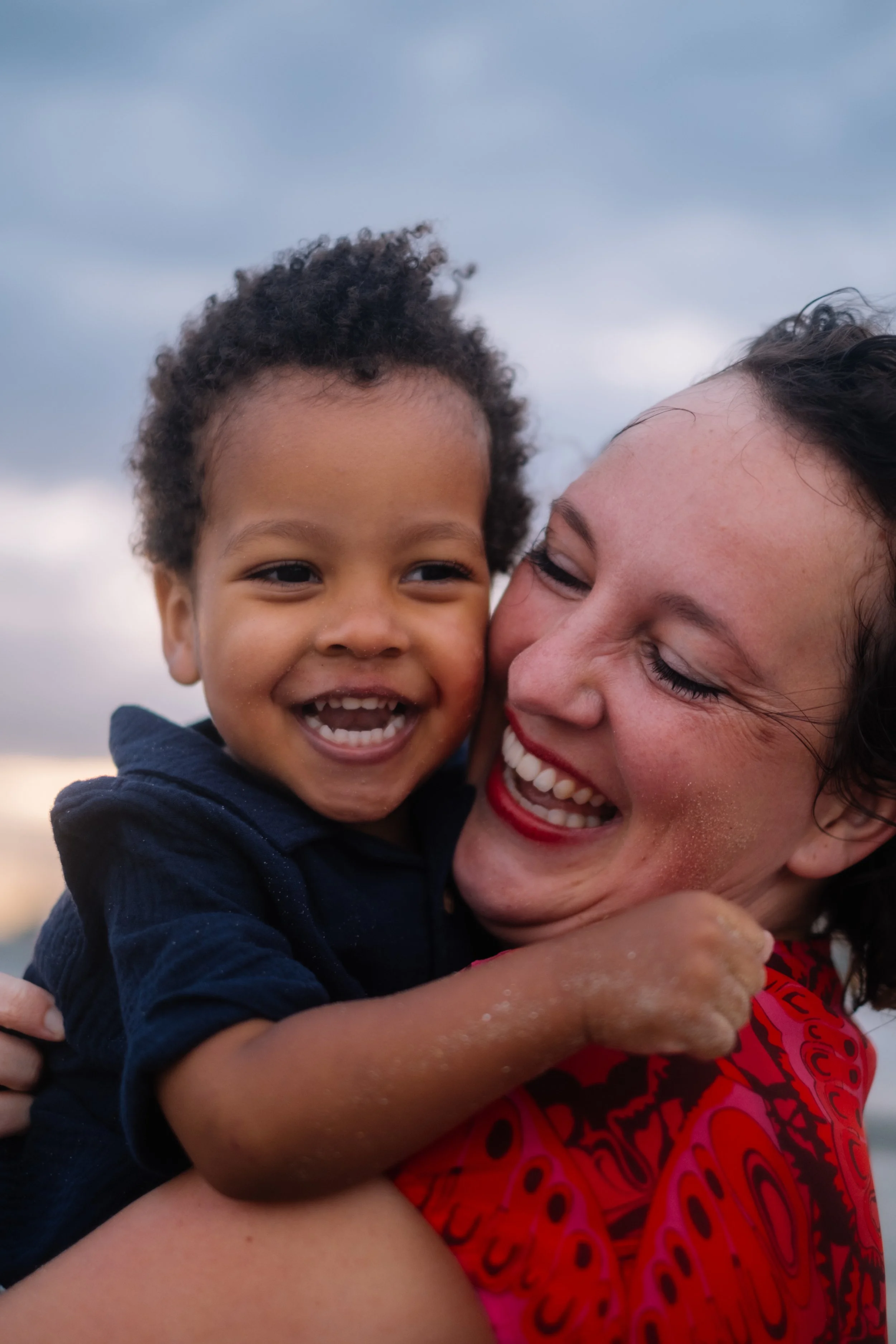 A woman and a young child are smiling and hugging. The woman has short dark hair and red lipstick, while the child has curly hair. The background shows a cloudy sky.