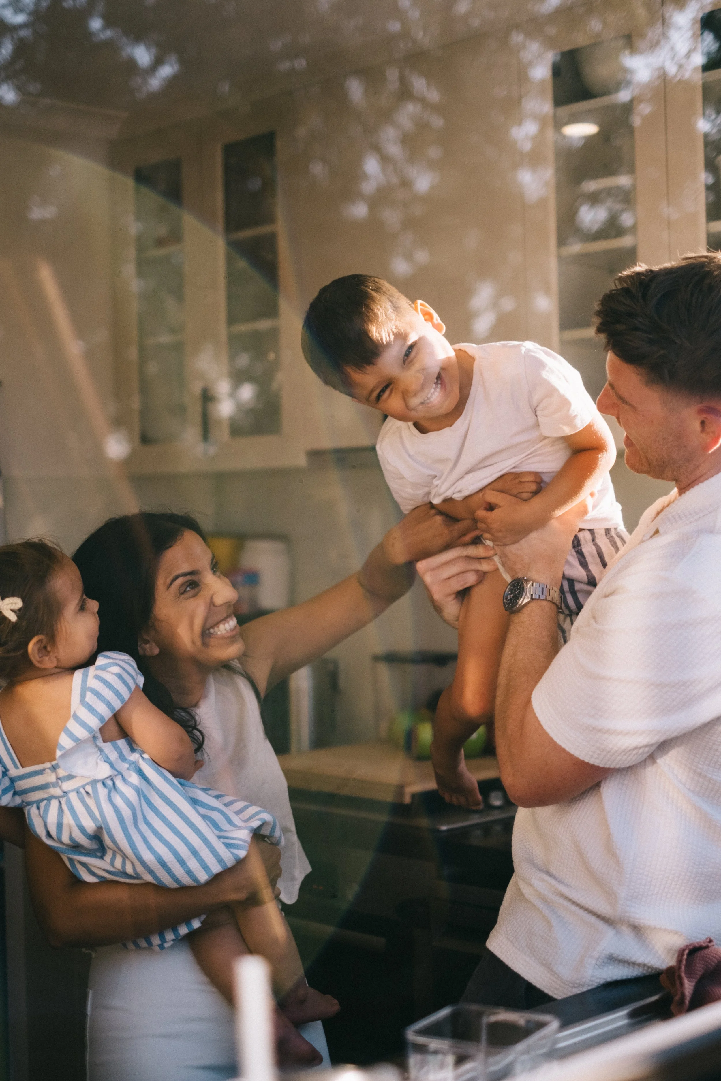 A happy family having fun and playing together inside their kitchen, seen through a glass window.