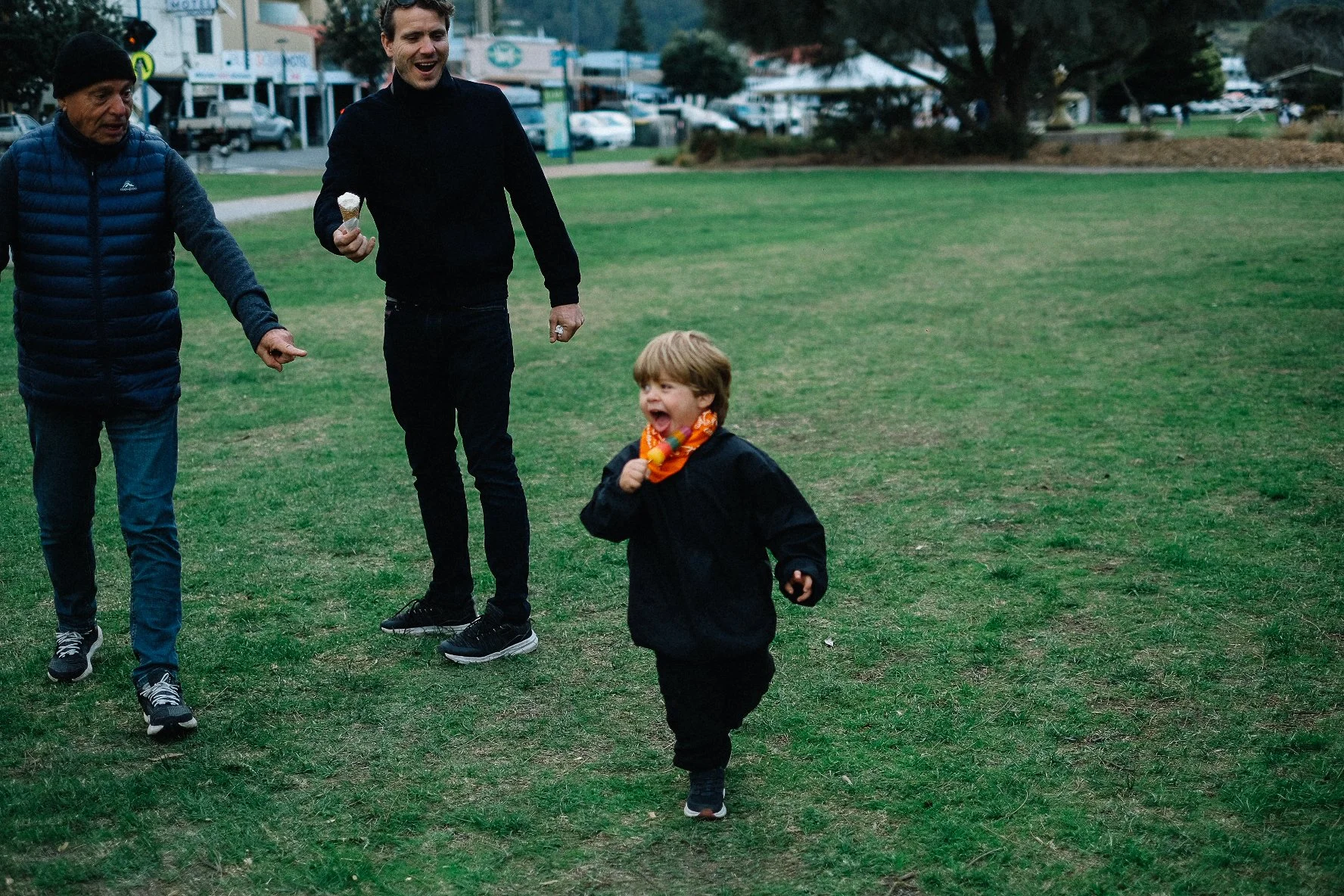 Three people, two adults and one child, walk and run across a grassy park field in the daytime, laughing and enjoying themselves, with buildings and trees in the background.